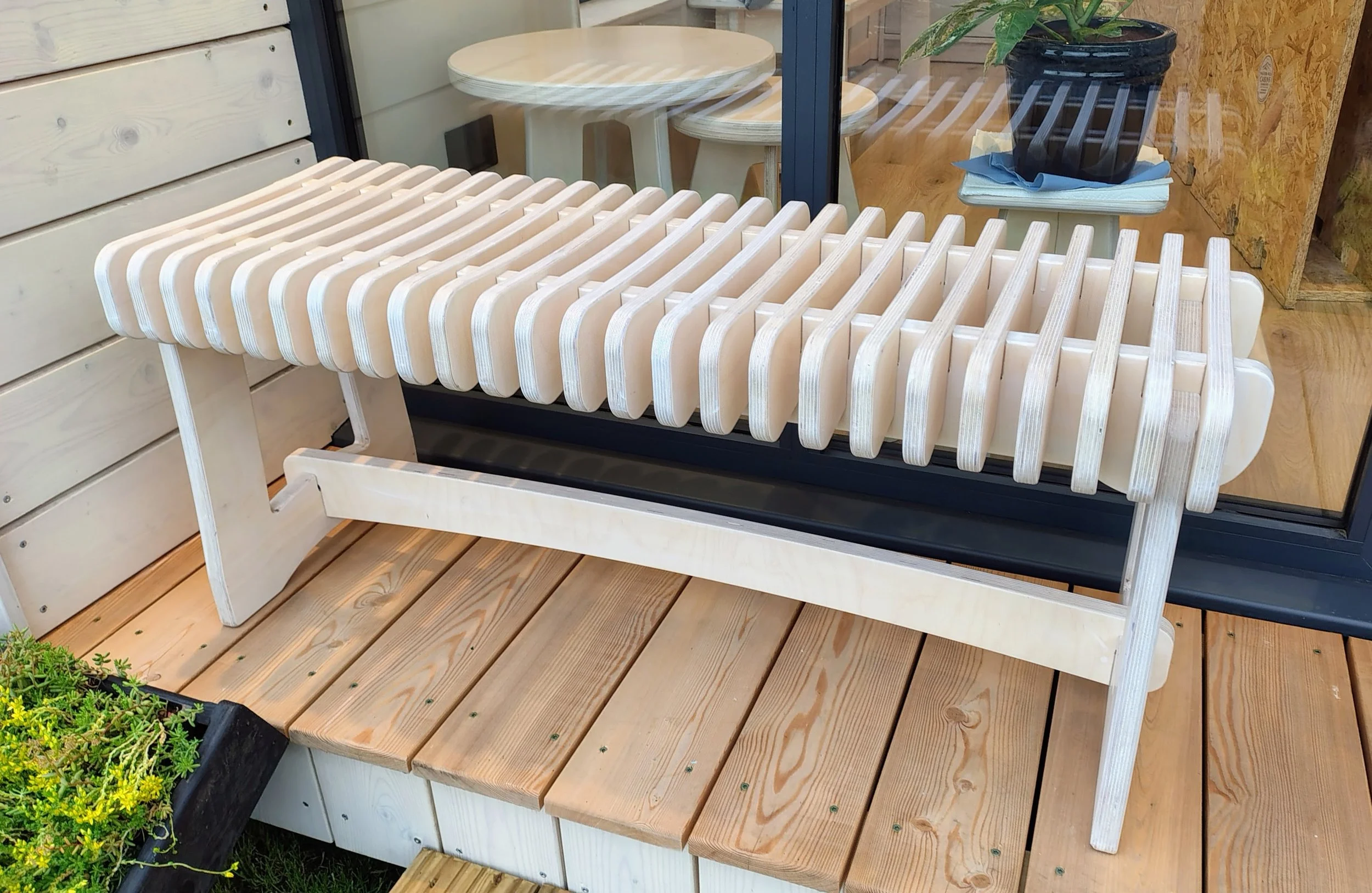 View of a wooden slat outdoor bench with a black decorative plant pot nearby, on a wooden deck with a wooden wall and glass window.
