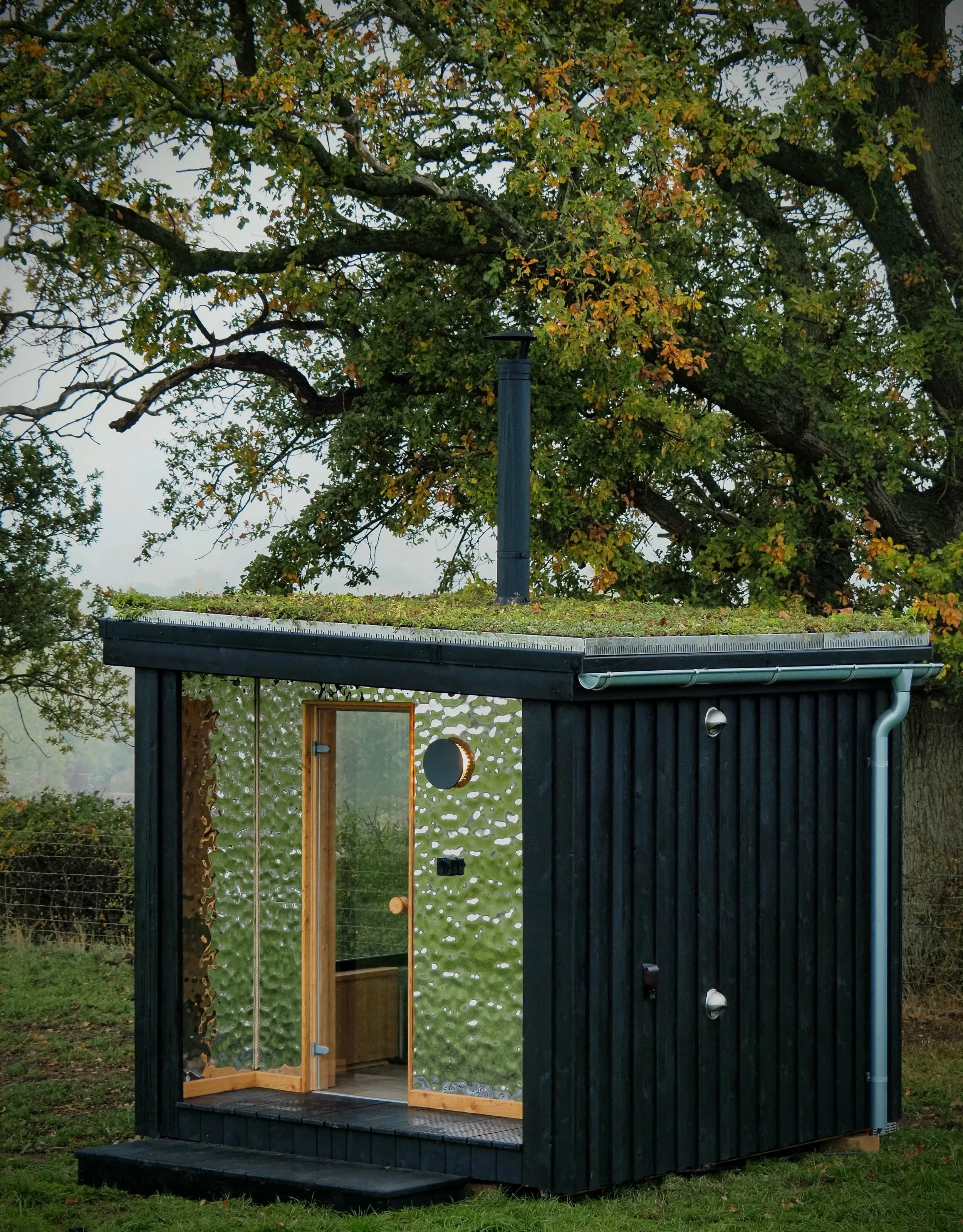 Small black outdoor sauna with a glass door and interior wooden bench, set on a grassy area with trees in the background.