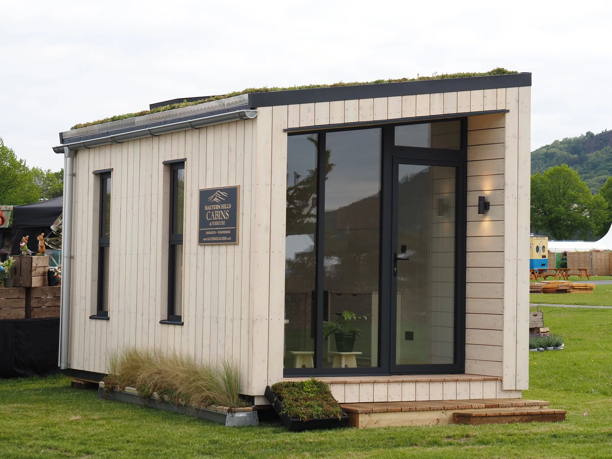 A small modern cabin with vertical light-colored wood siding and black window and door frames, sitting on a grassy area with green trees and hills in the background.