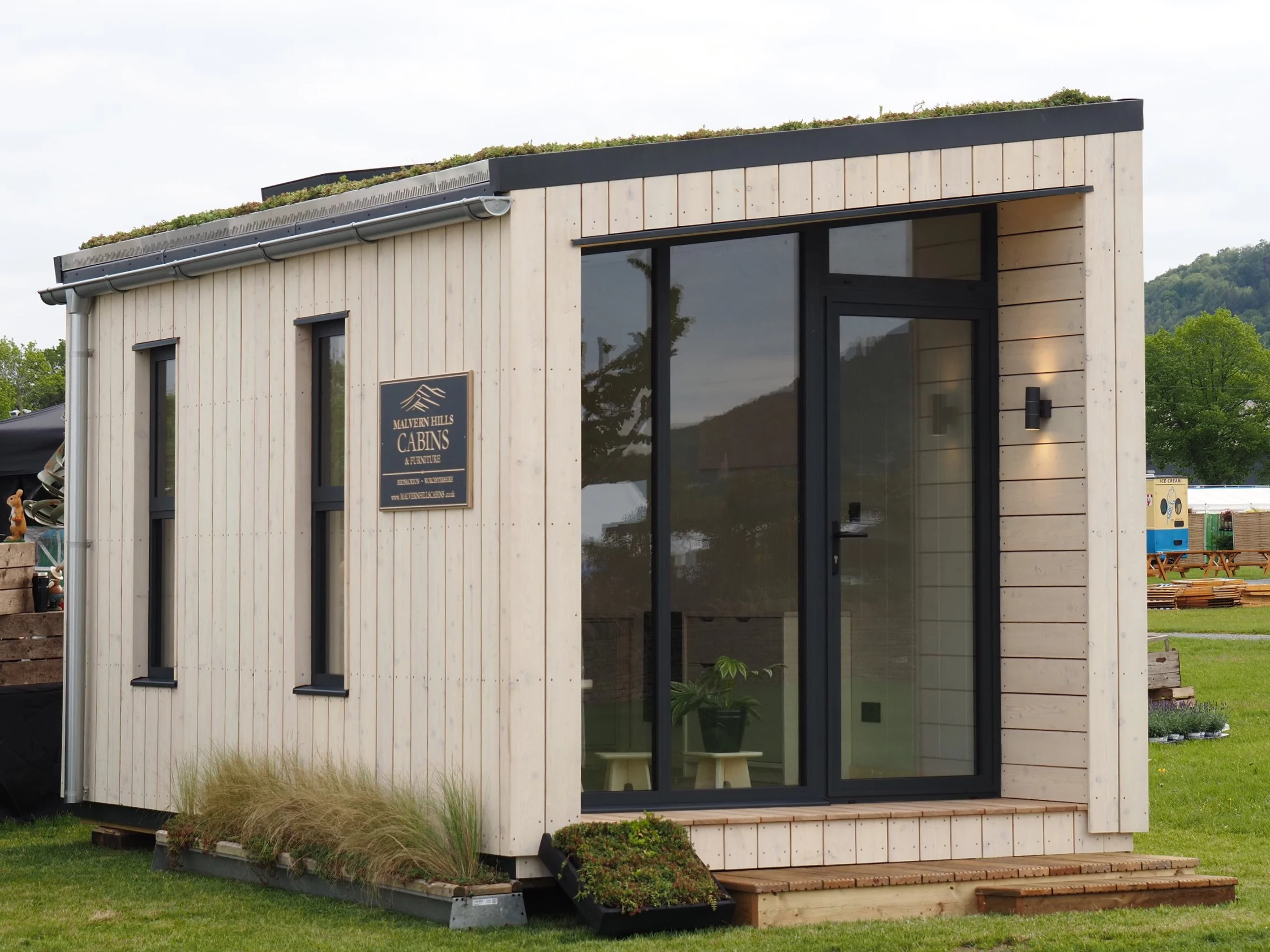 A small modern building with a wooden exterior, large glass doors, and windows, surrounded by green grass and trees, with a mountain in the background. There is a sign that reads 'Malvern Hills Cabins & Furniture.'