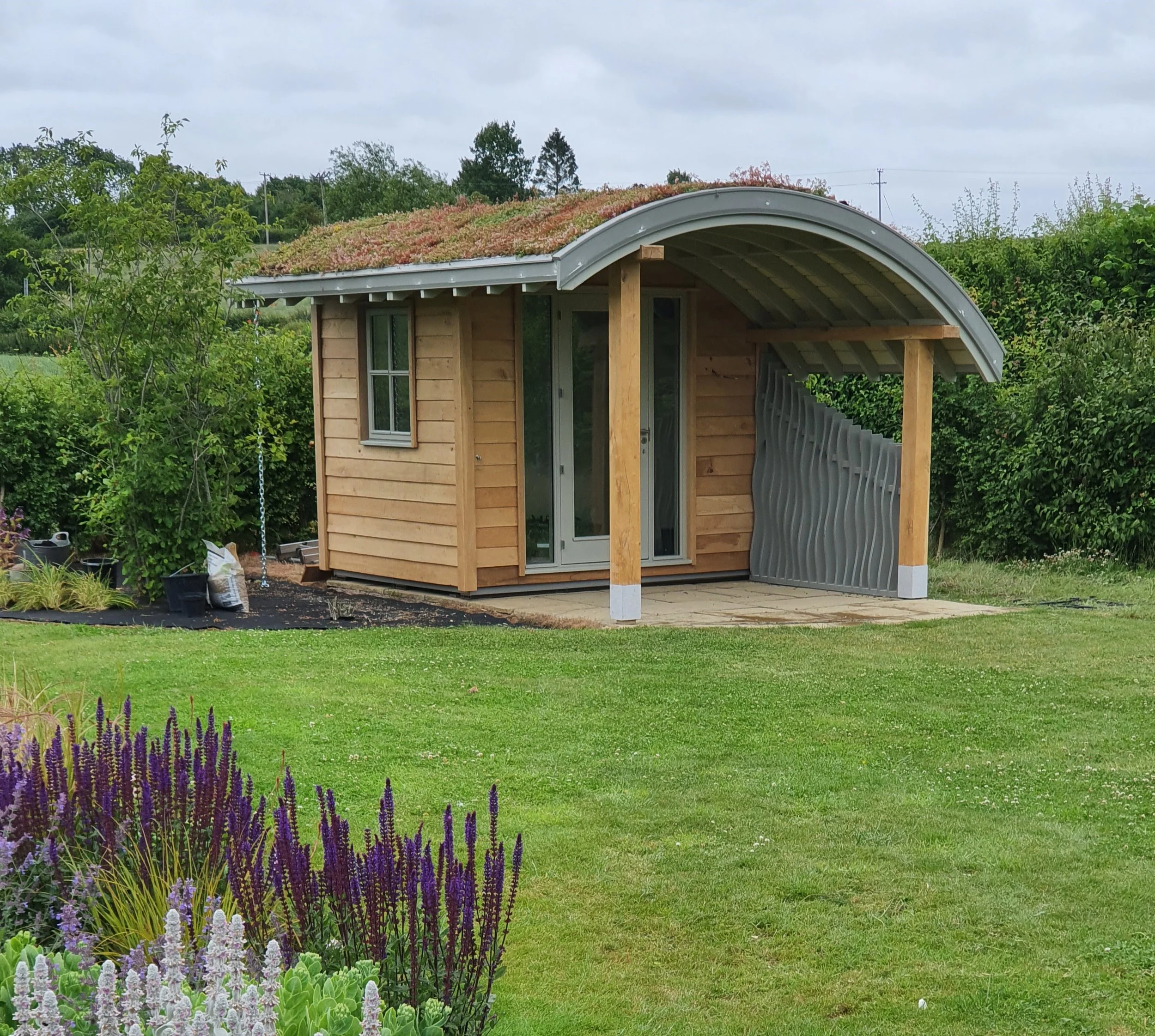 A small wooden garden shed with a curved metal roof covered in green and brown plants, surrounded by green trees and purple flowers in the foreground under an overcast sky.