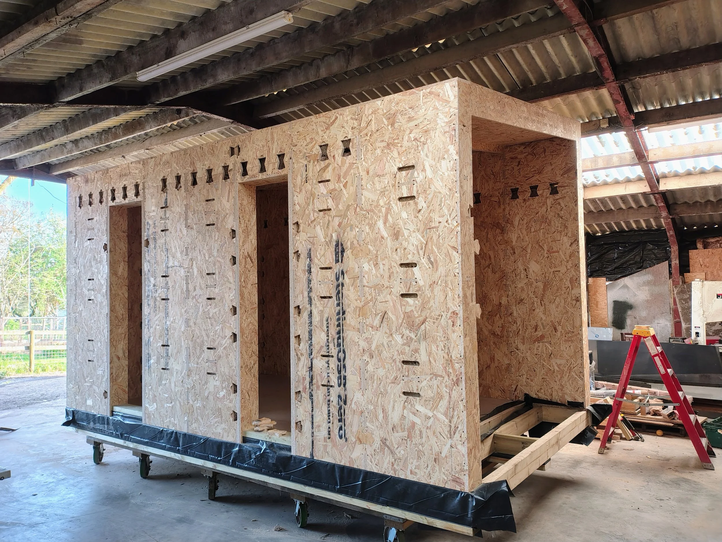 Construction site with a wooden structure built from OSB boards inside a building with a corrugated metal roof and exposed beams. There is a ladder, tools, and construction materials around.