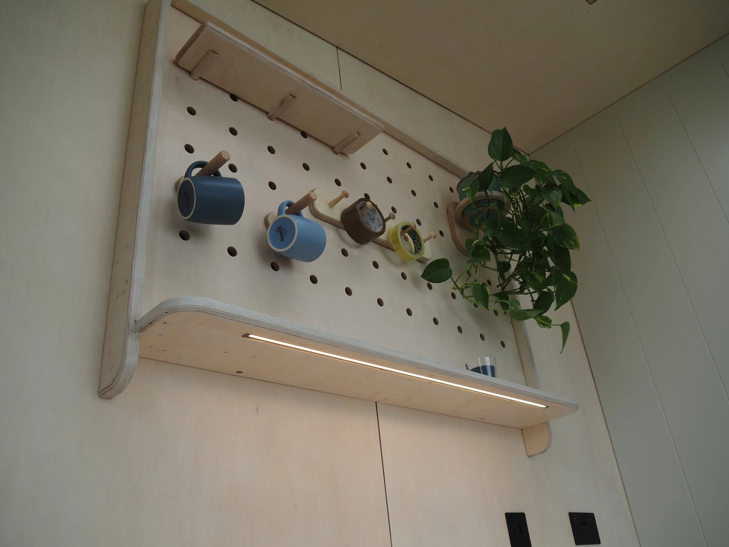 A wooden pegboard with various small wooden shelves and hooks, decorated with two blue coffee mugs and a potted plant with green leaves.