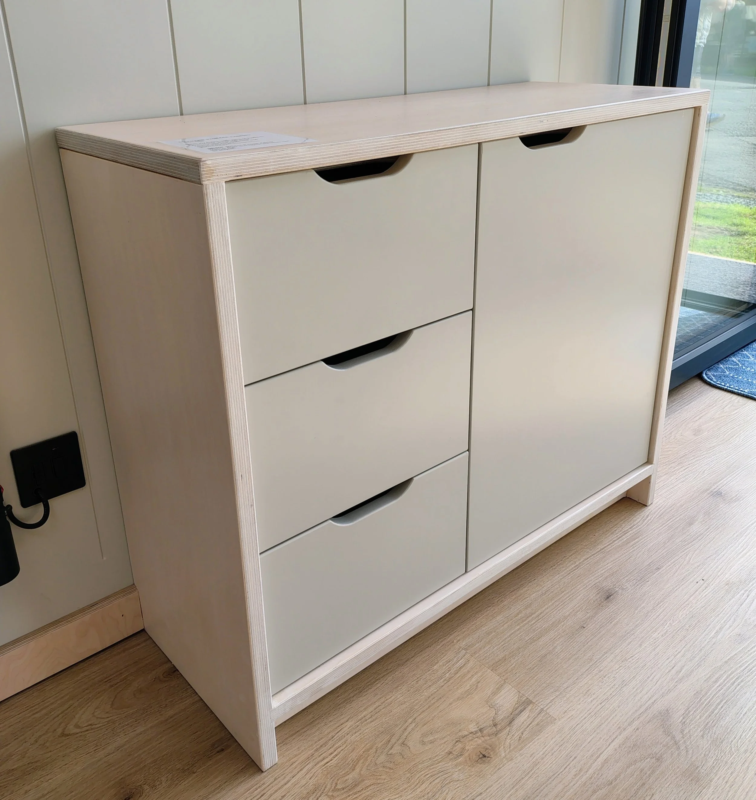 Unfinished wooden cabinet with four drawers and a light wood countertop, positioned against a wall with vertical panels, located next to a glass door or window.