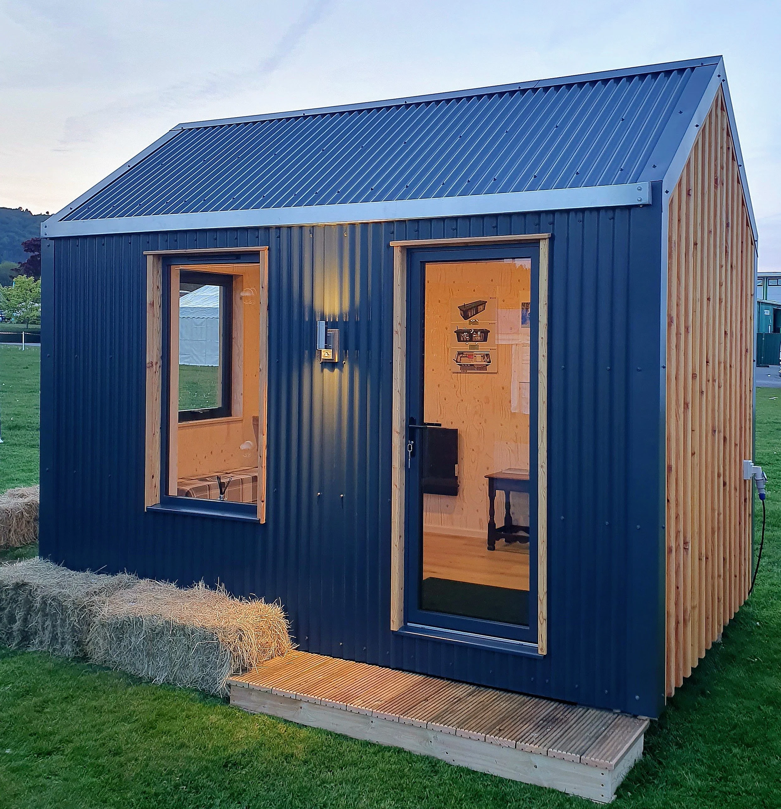 Small house with blue corrugated metal roof and dark blue siding, large front window, wooden trim, and a wooden deck in front, in a grassy yard with hay bales to the left.