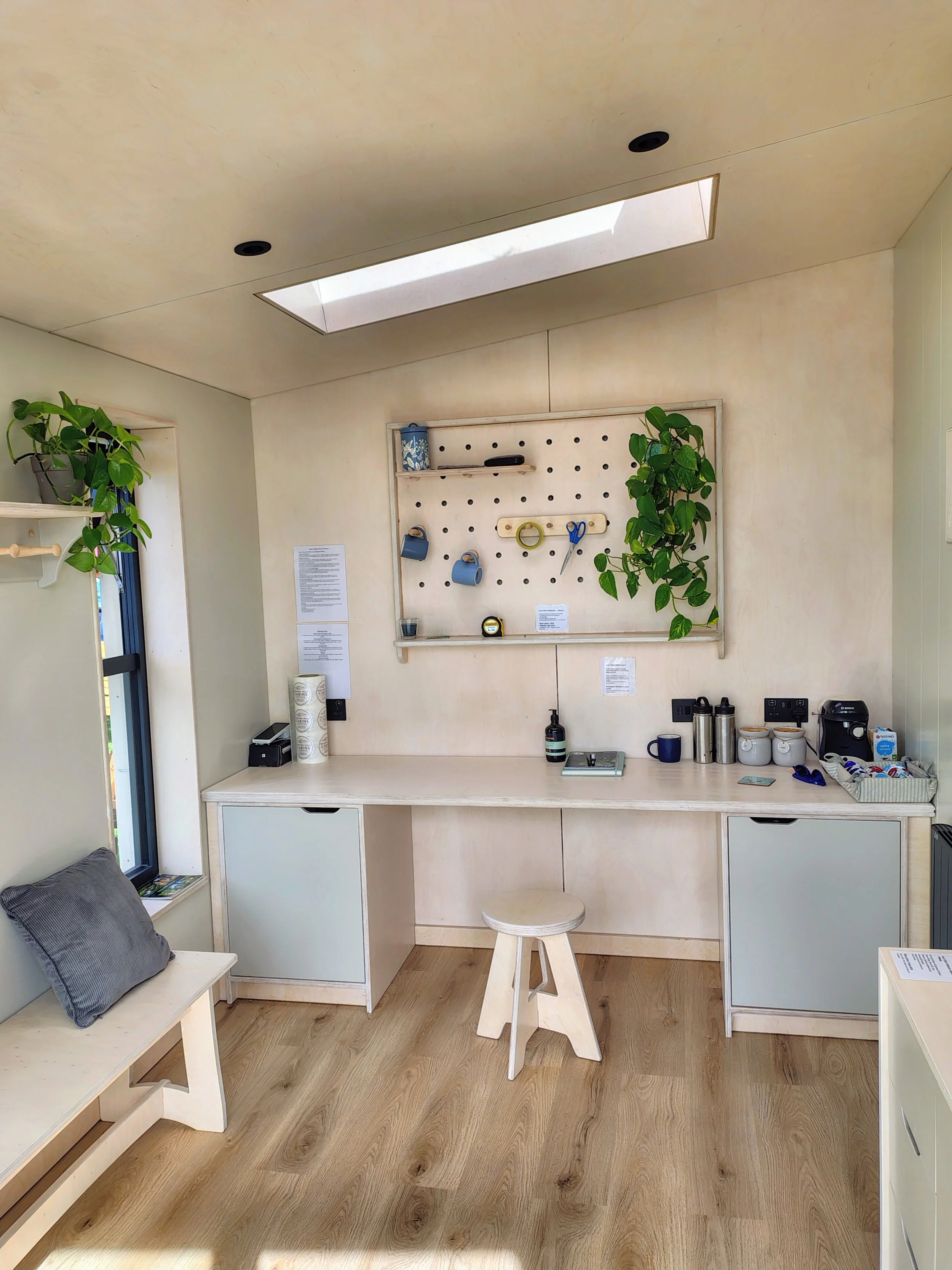 A small kitchen corner with a white countertop desk, a white stool, and a white table with a gray pillow. The wall has a pegboard with small items and plants. There are two plants, one on each side of the pegboard, and a skylight above.
