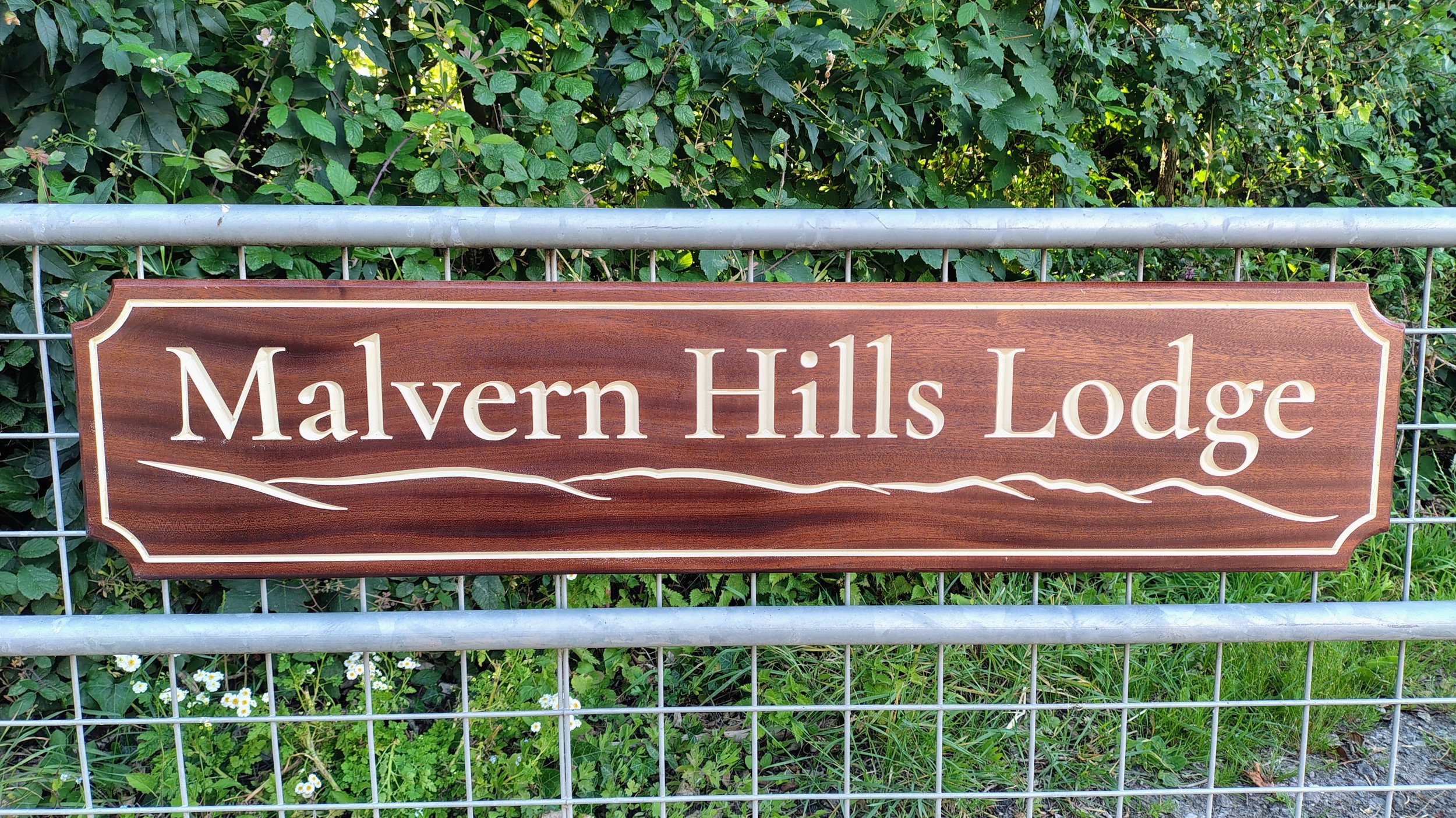 Wooden sign that reads 'Mavern Hills Lodge' with a mountain and wavy line illustration, mounted on a metal fence, with lush green foliage and small white flowers in the background.