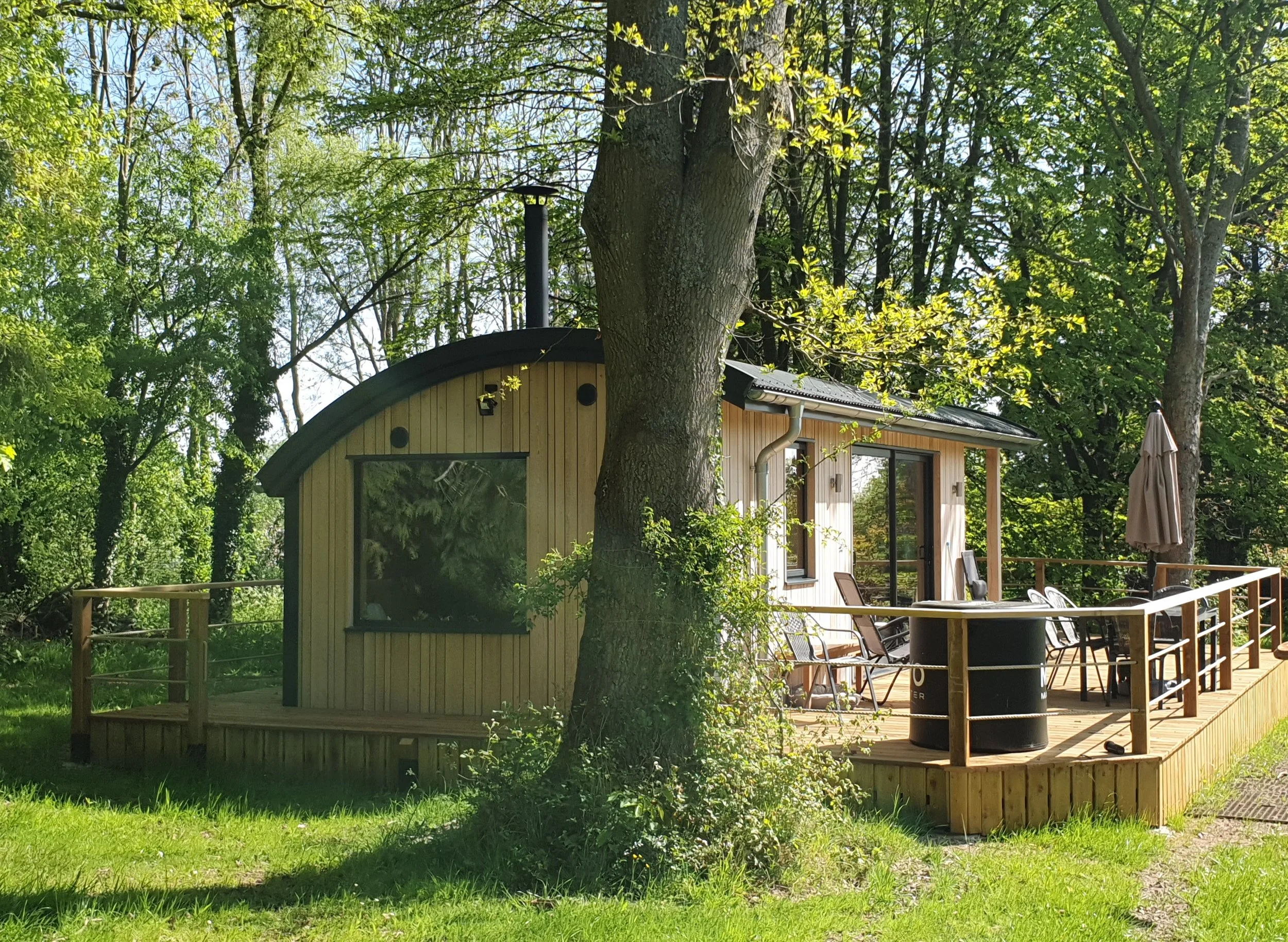 A wooden cabin with large windows and a sliding glass door, surrounded by green trees and a well-maintained lawn. There is a deck with outdoor chairs, a table, and an umbrella, and a black trash bin nearby.