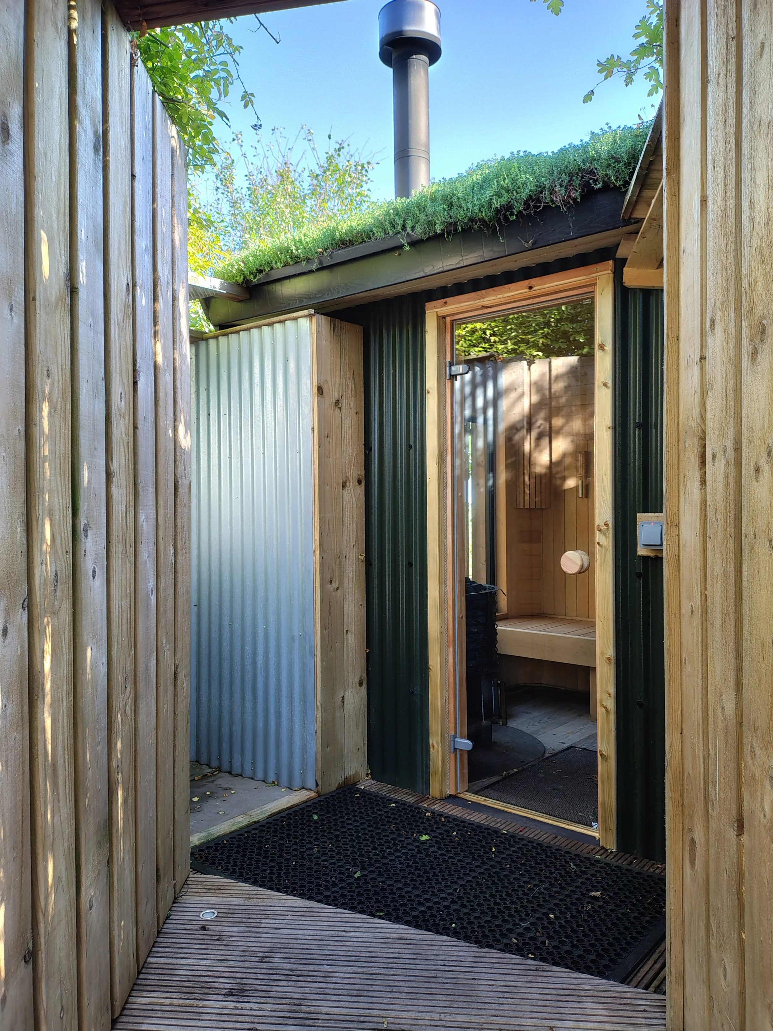 View of an outdoor wooden entrance with a metal door, a wooden interior doorway, and a black rubber mat on the ground. The roof is covered with greenery, and a chimney extends into a blue sky. There are trees and foliage visible around the structure.