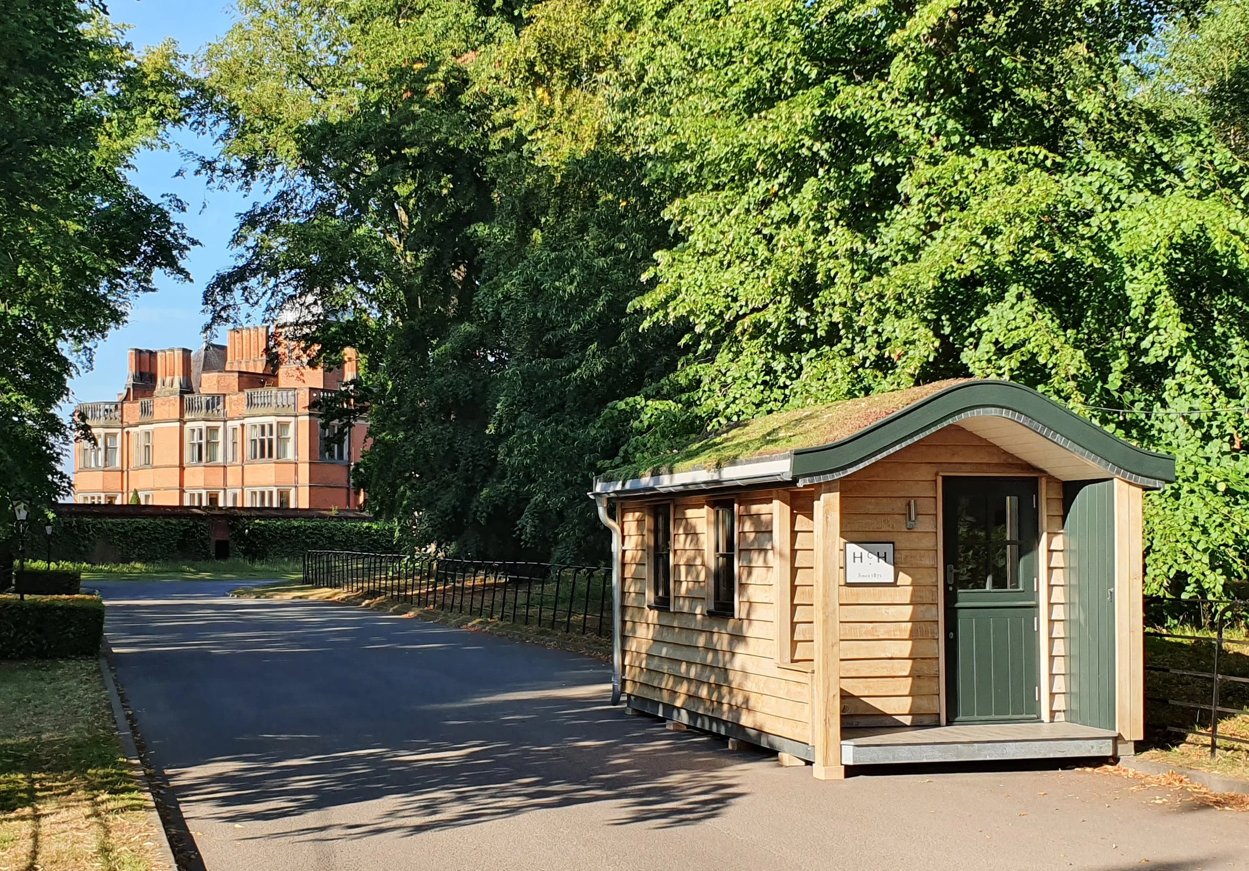 A small wooden structure with a green door and window, set on a paved area, surrounded by trees and greenery, with a large brick building in the background.