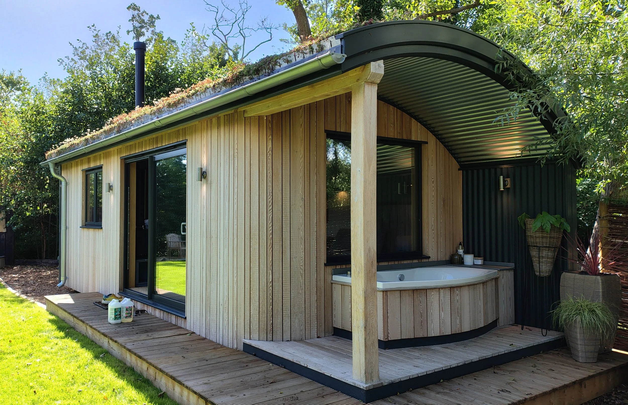 A modern backyard house with vertical wooden siding, large glass doors, a curved metal roof, and an outdoor bathing area with a round tub, surrounded by greenery and trees.