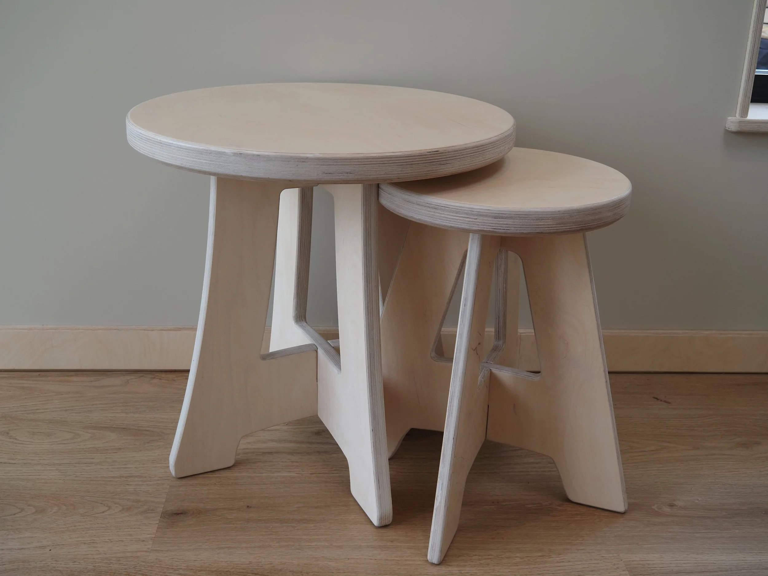 Three wooden nesting stools with rounded tops and angled legs, arranged on a wooden floor against a light-colored wall.