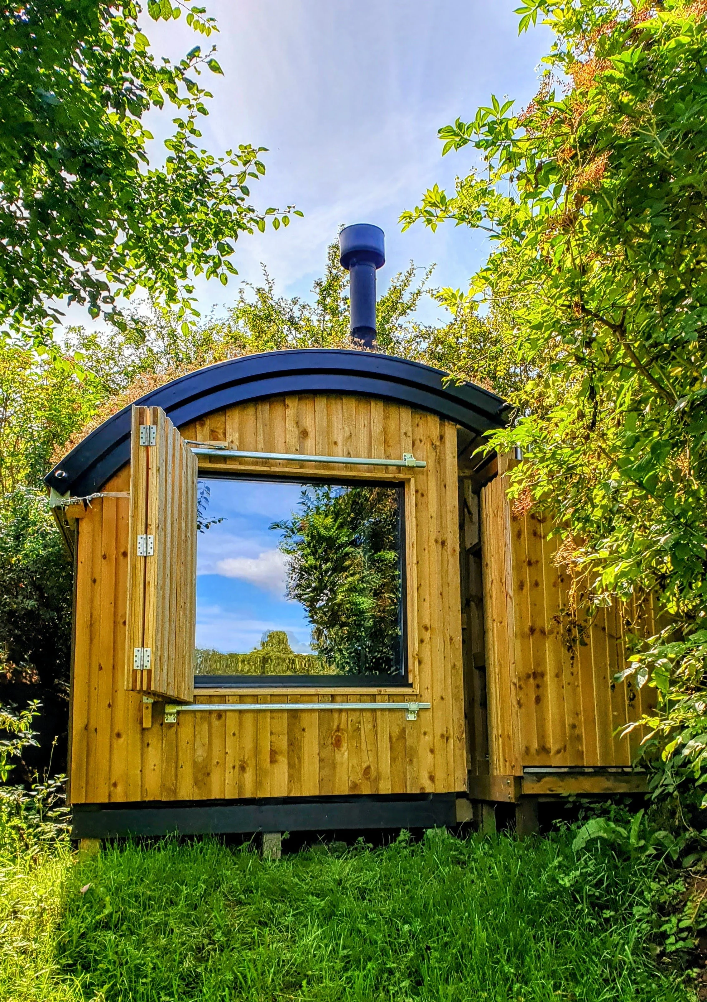 A small wooden house with an arched roof and a large window that reflects the sky and trees, surrounded by lush green bushes and grass.