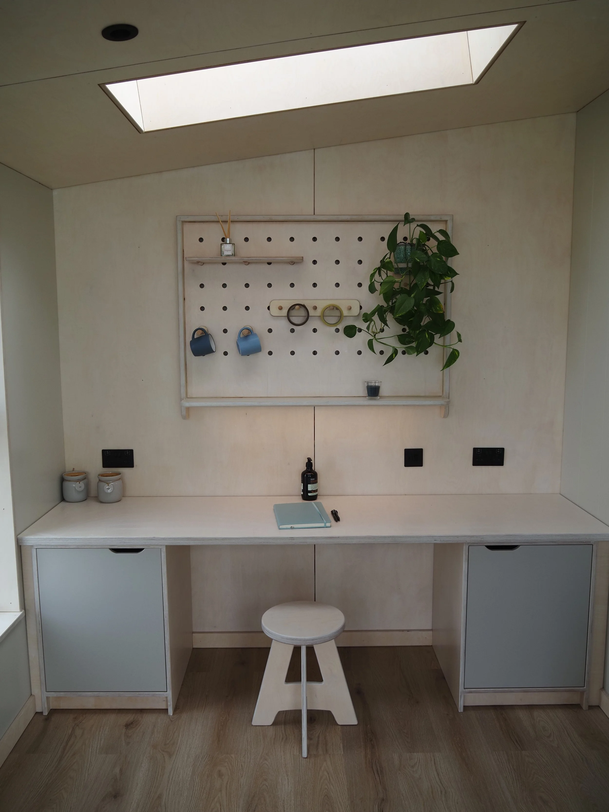 A white workspace with a small wooden stool, two white cabinets, a desk, a pegboard with hanging mugs, a potted plant, a notebook, pens, and a bottle of sanitizer. There is a skylight in the ceiling.