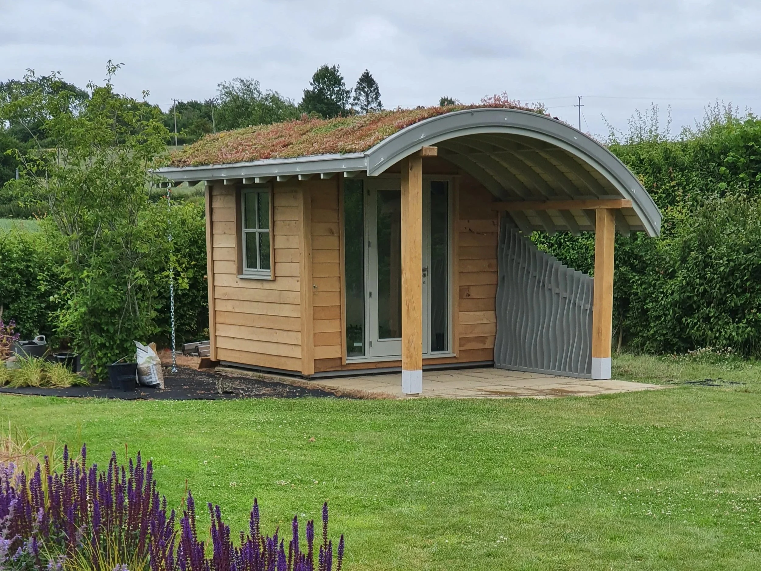 A small wooden shed with a curved metal roof, surrounded by green trees and plants, with purple flowers in the foreground, and an overcast sky.