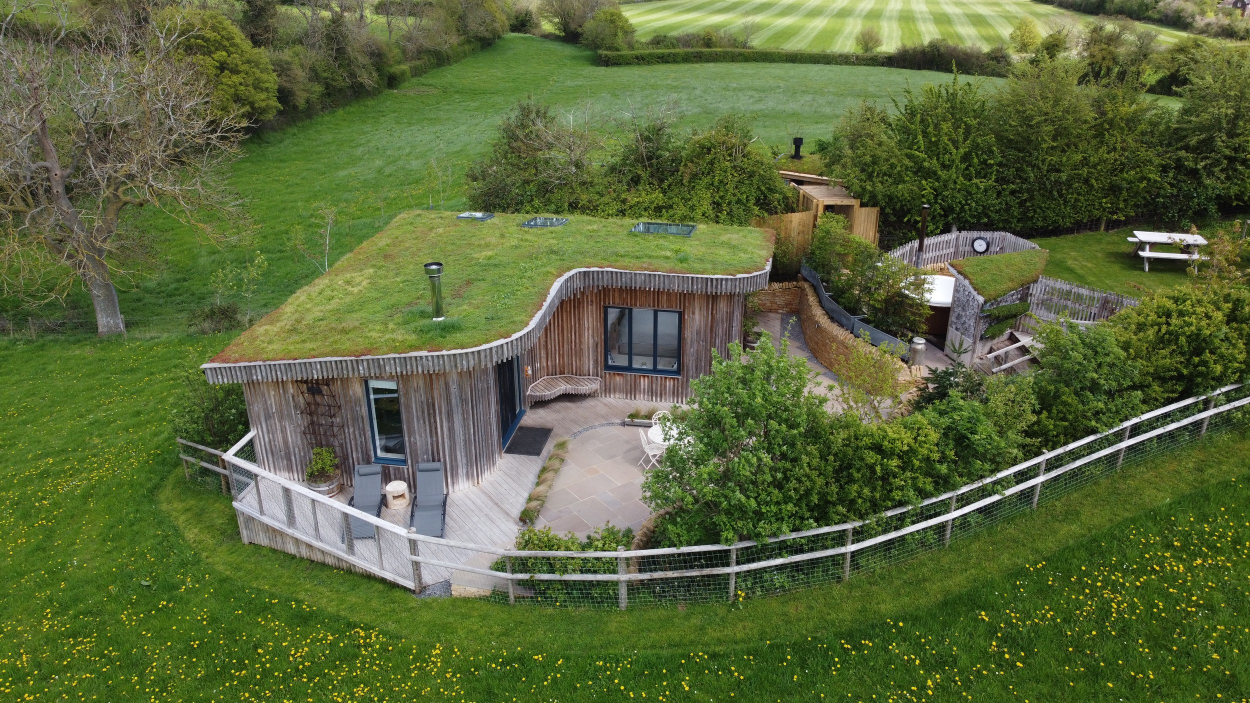 A modern house with a curved, green roof and wooden exterior walls, surrounded by lush greenery and a fenced yard, with outdoor seating and a patio.