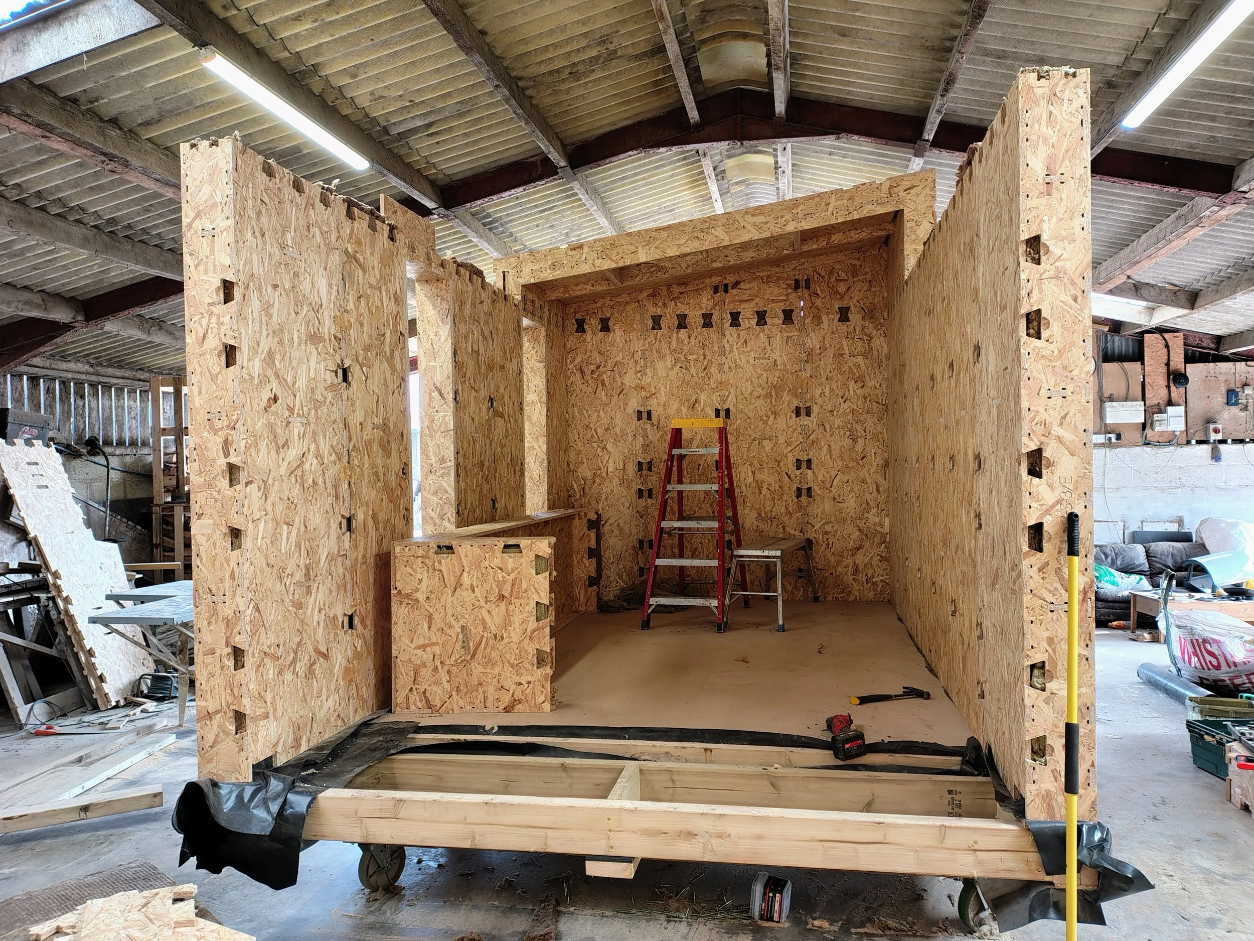 Interior of a building under construction with wooden framing, studs, and plywood walls, along with construction tools and materials scattered around.
