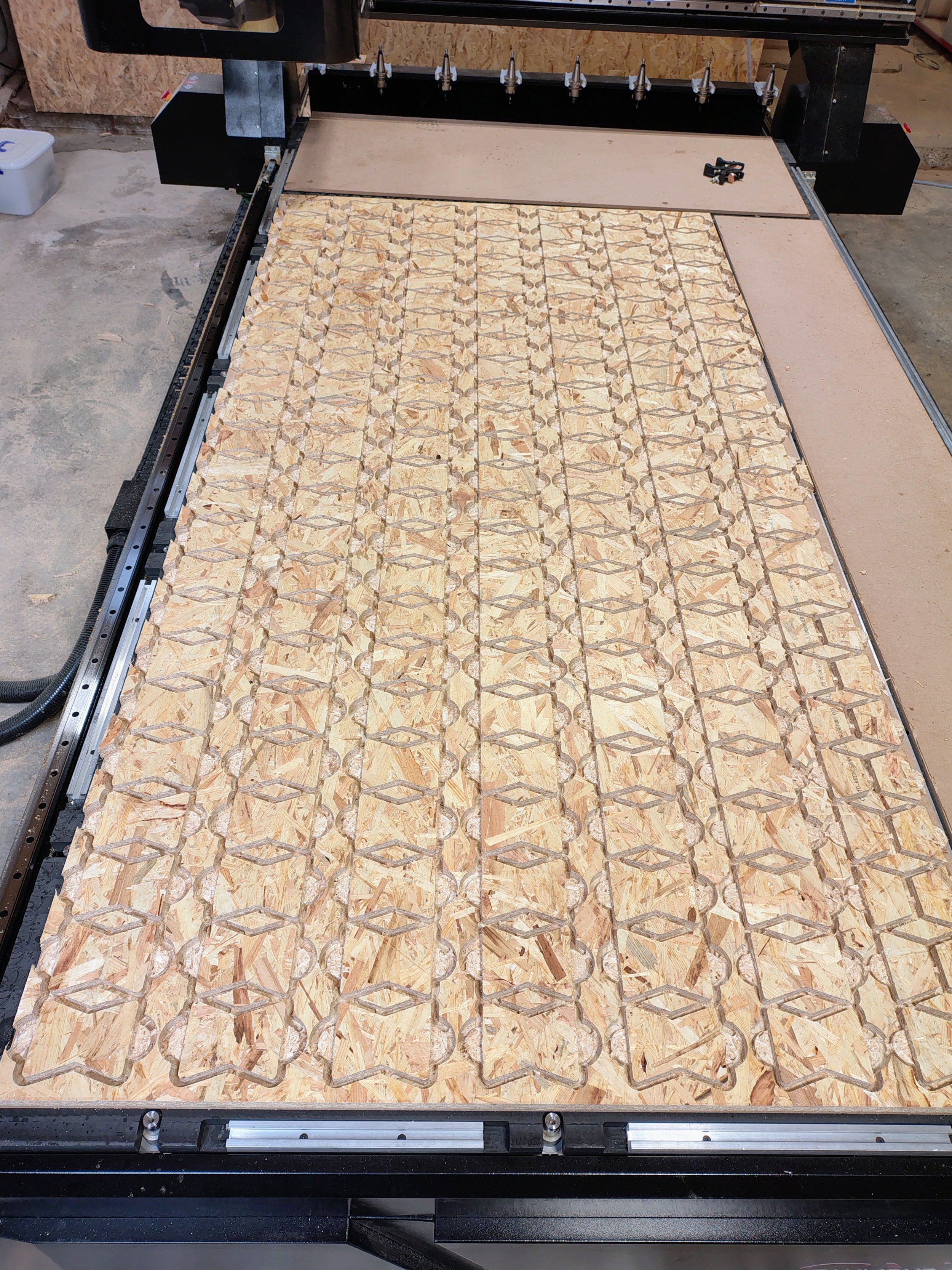 A CNC machine cutting a large sheet of oriented strand board (OSB) into a geometric pattern. The machine has a black frame and a flat surface with a section covered in patterned cuts, and the workspace appears to be in a workshop or garage.
