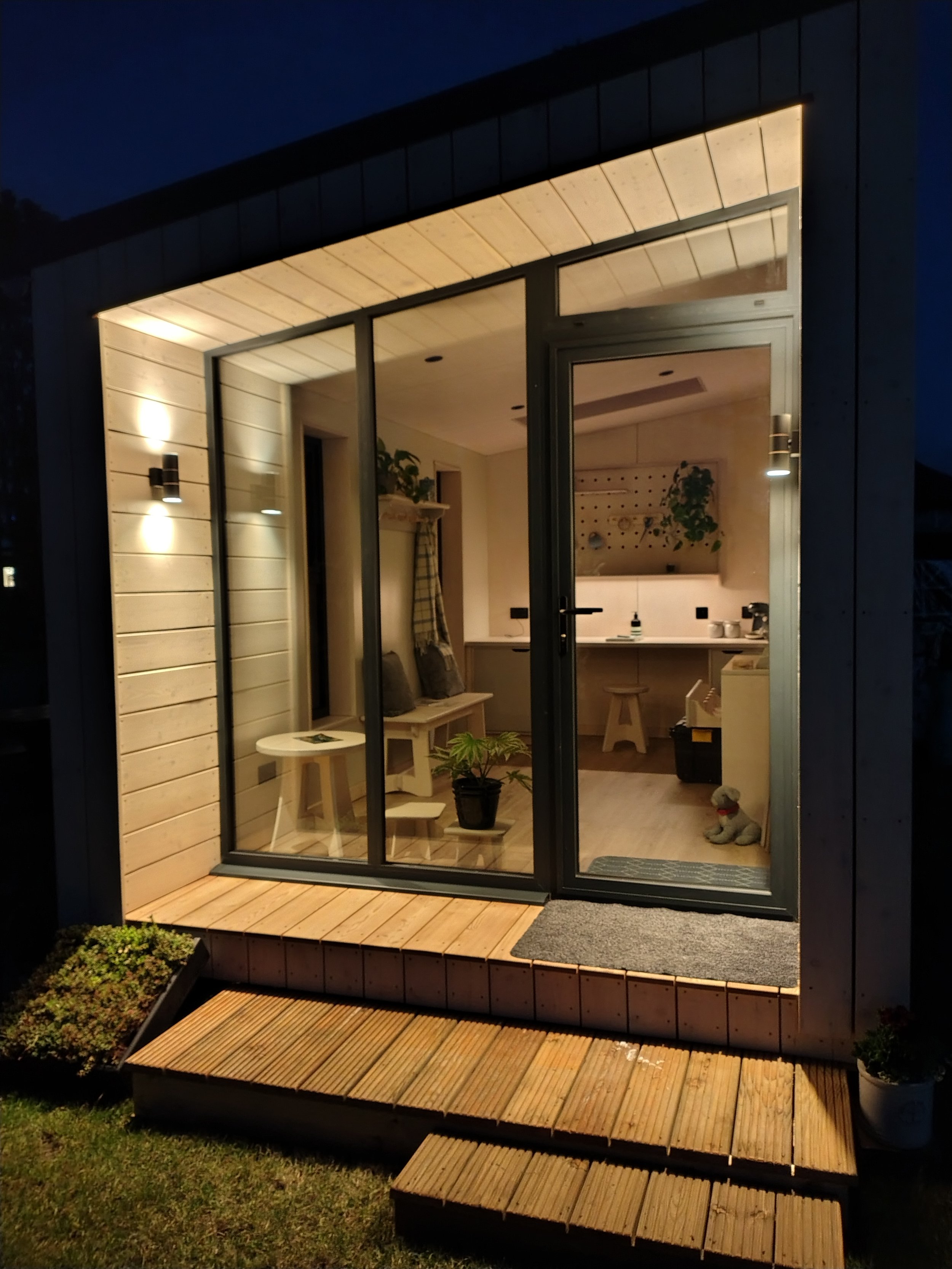 Nighttime view of a small modern house with large glass windows and a wooden deck. The interior is warmly lit, showing a dining and kitchen area with plants and furniture. A small dog figure is sitting near the door, and a potted plant is on the deck outside.