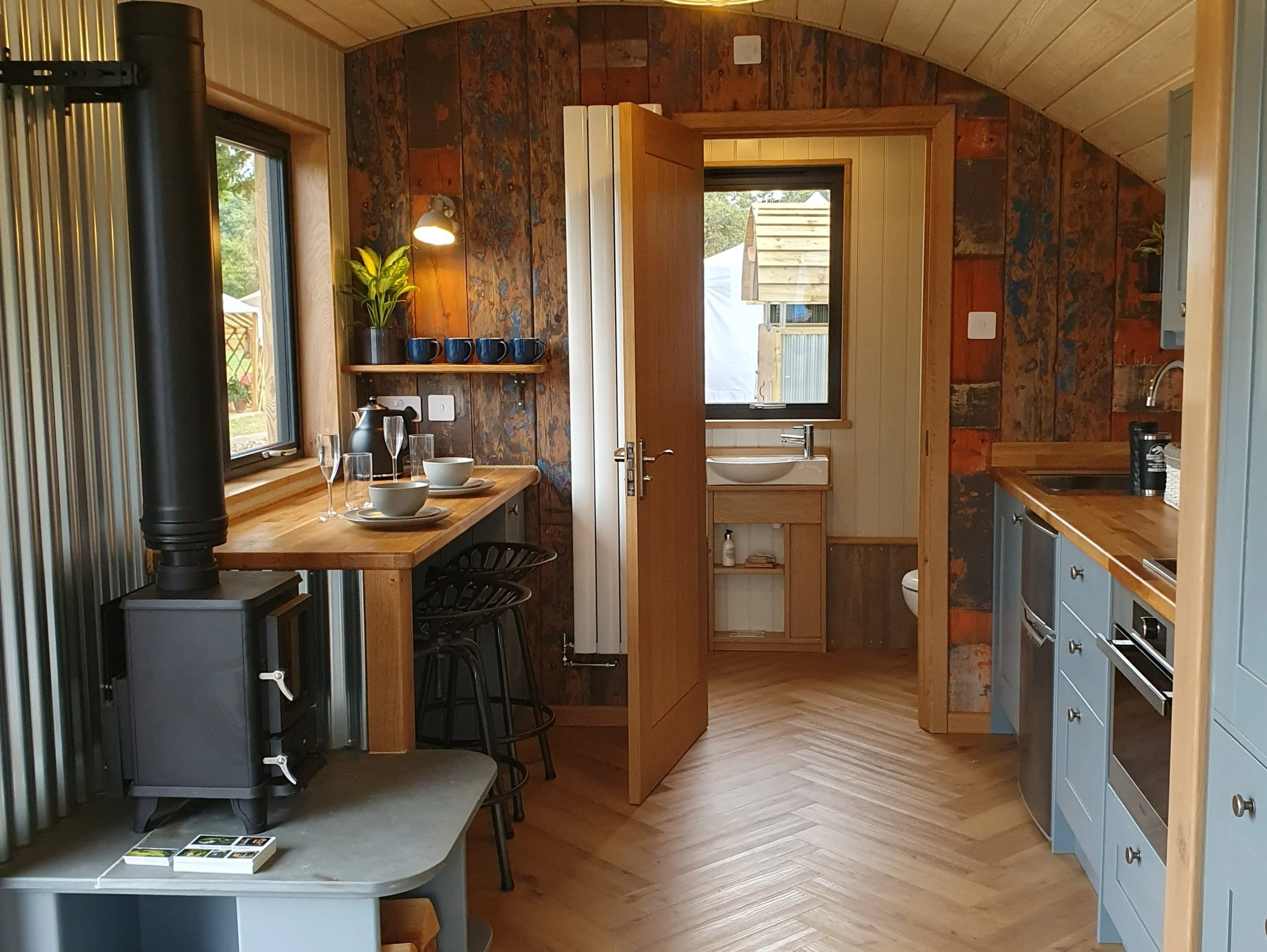 Cozy kitchen with wooden walls, a small dining table set with bowls, glasses, and a coffee pot, black bar stools, and a wood stove. Two windows display a backyard with trees and a garden, and a door opens to an outdoor space with a wooden fence.