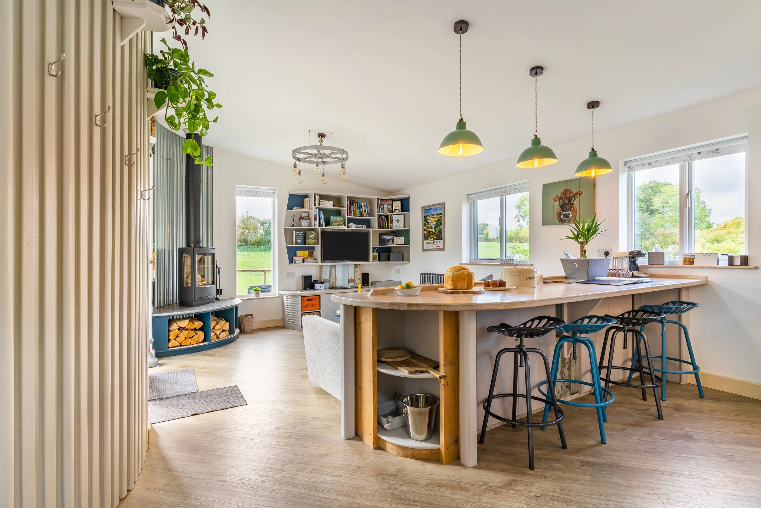 A cozy kitchen and living space with a large island, black bar stools, several windows, a bookshelf, and a wood-burning stove, decorated with plants and artwork, with a view of greenery outside.