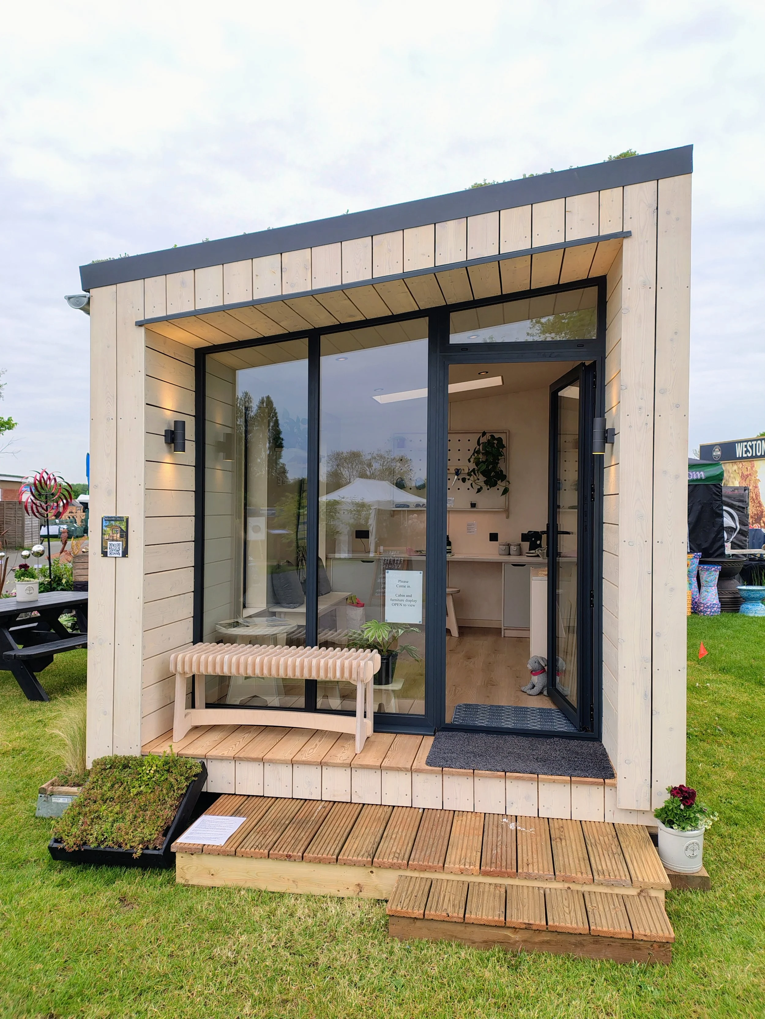 Small modern wooden house with large front glass doors, black framing, a wooden bench outside, and potted plants, situated on a grassy area during daytime.