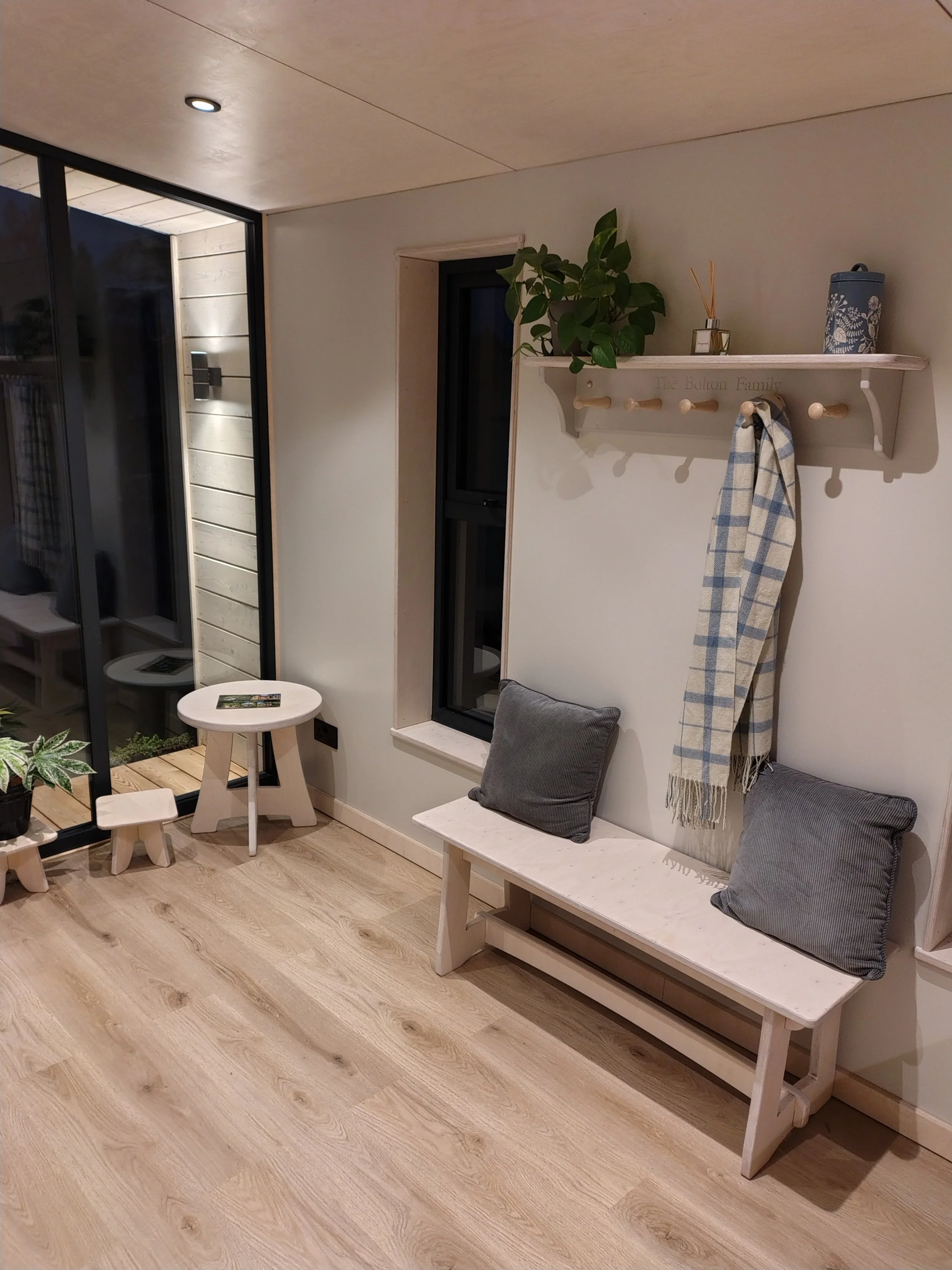 Living room with wooden floor, white bench with gray cushions, wall shelf with plants and candles, small round white side table, and sliding glass door leading outside.