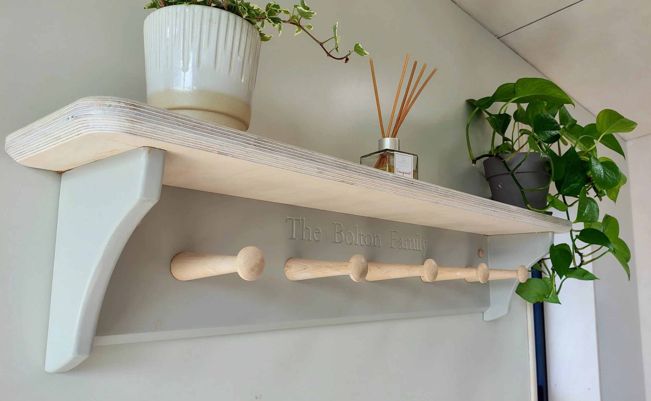 A decorative wall shelf with a potted plant, a reed diffuser, and hanging hooks with a sign reading 'The Bolton Family' beneath.