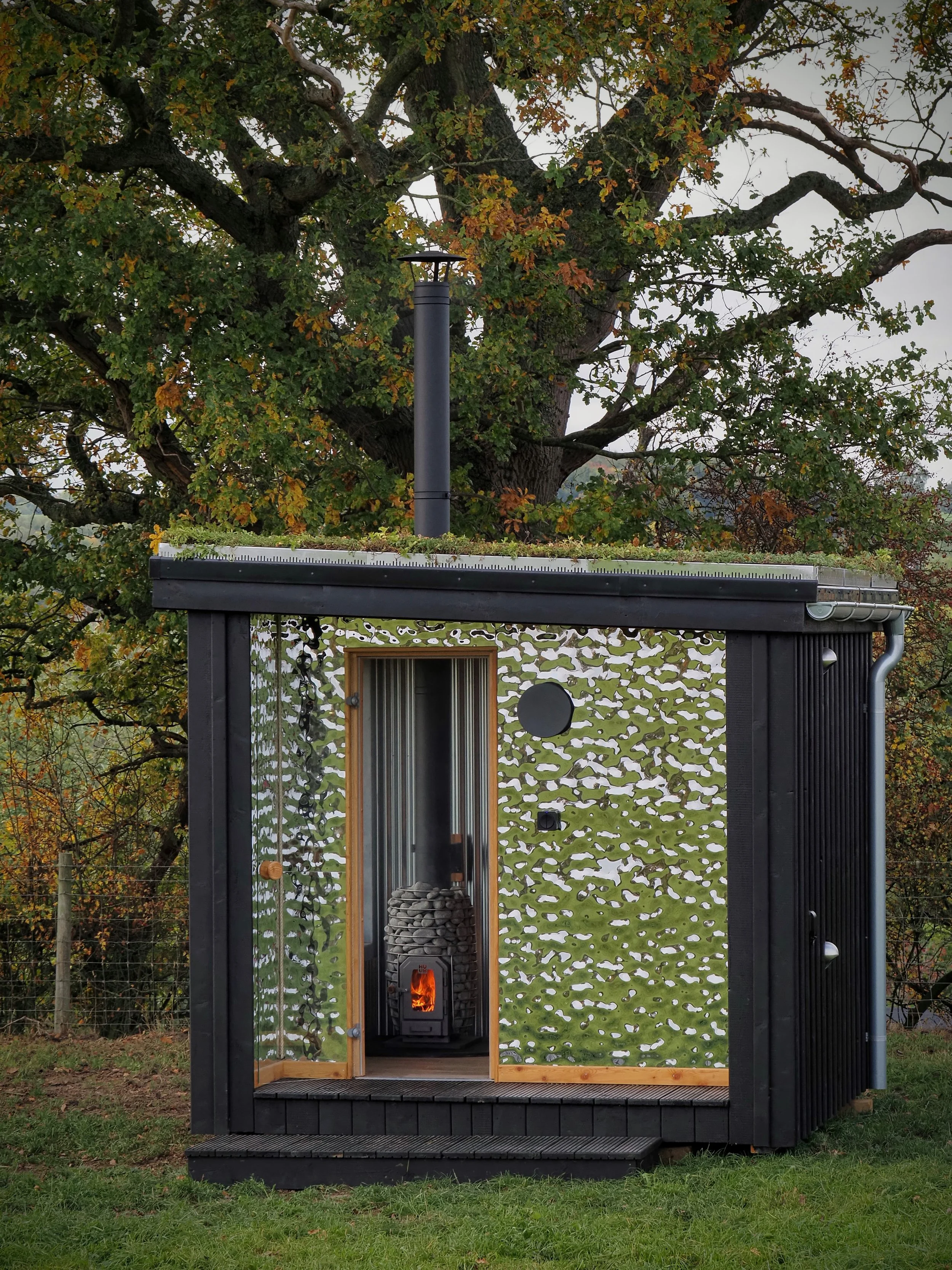 Small black cabin with a wood stove and a glass door, surrounded by a patterned metal frame with a camouflage design, set outdoors on grass with trees in the background.