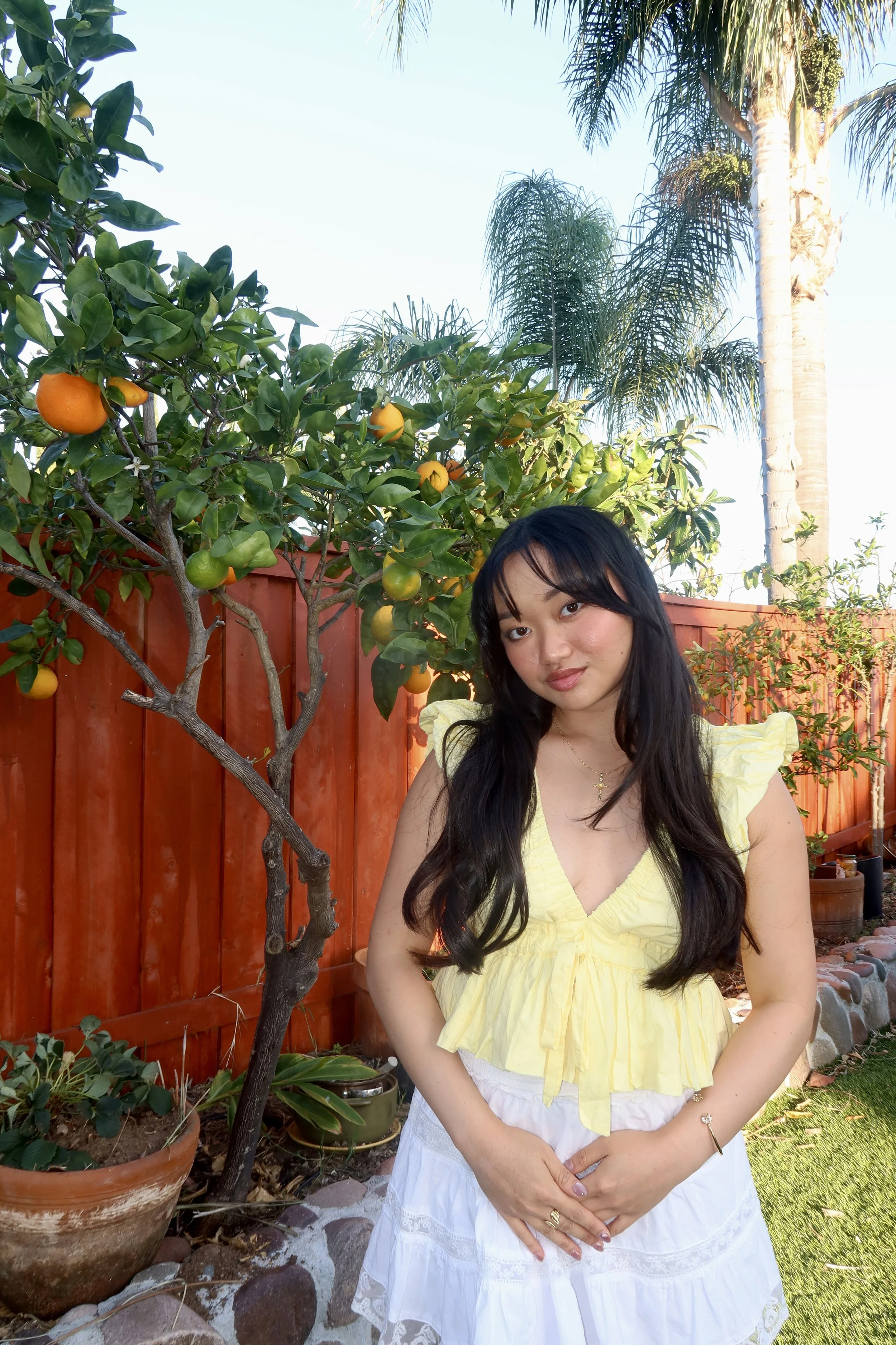 Young woman with long dark hair and a light yellow top standing in a backyard garden with trees, plants, and a red wooden fence in the background.