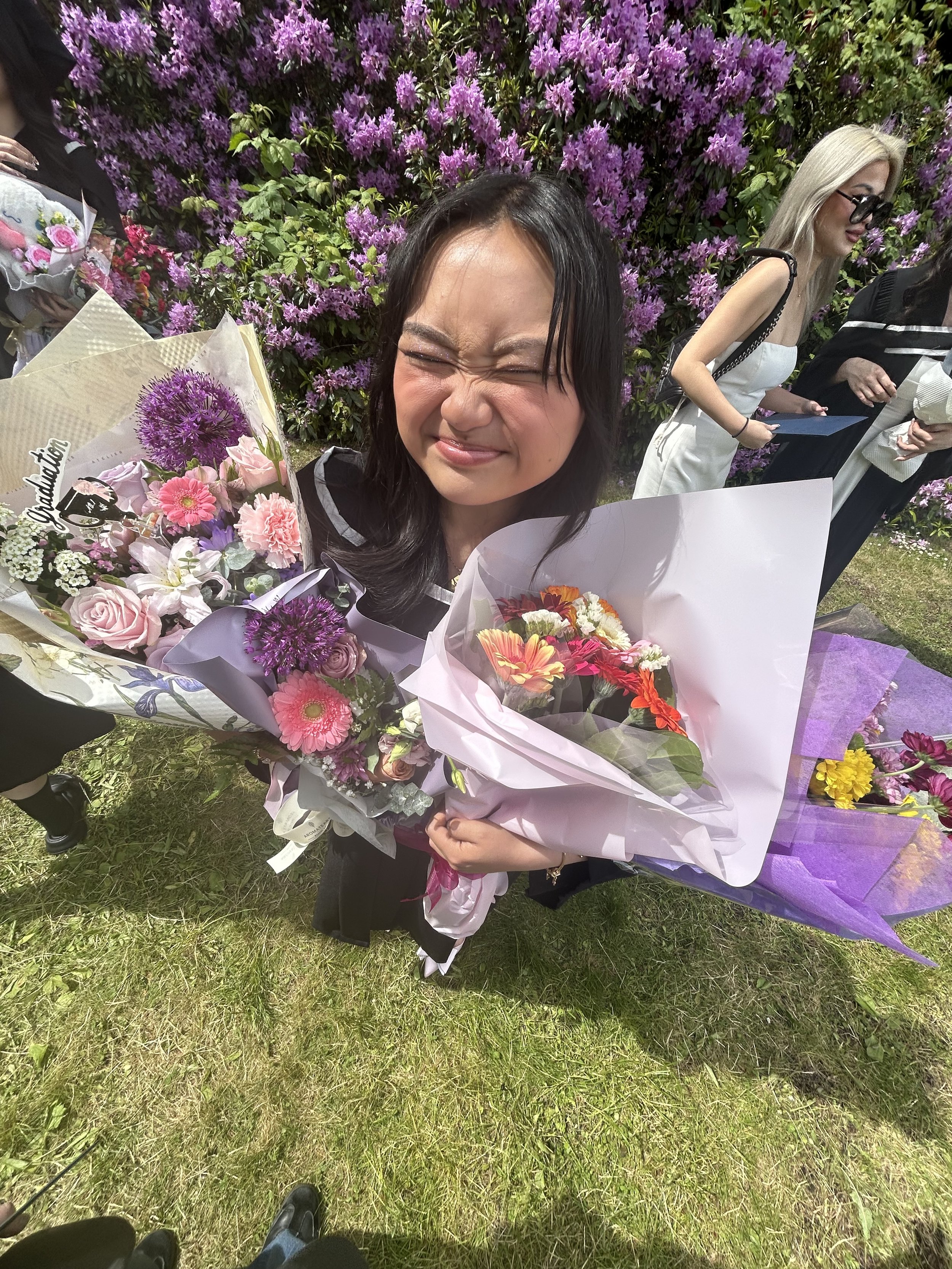 A woman with black hair and closed eyes holding a bouquet of colorful flowers, standing outdoors in front of a bush with purple flowers. Others are visible in the background, and the photo is taken from above, showing the grass and feet of people at the bottom.