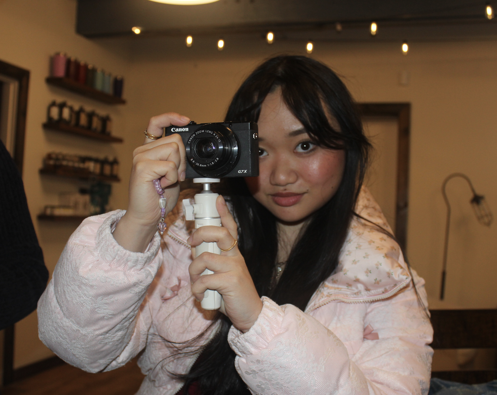 A young woman with long dark hair and a pink floral top holding a camera on a tripod, looking directly at the camera with a slight smile in a warmly lit indoor setting with string lights overhead and shelves with various items in the background.