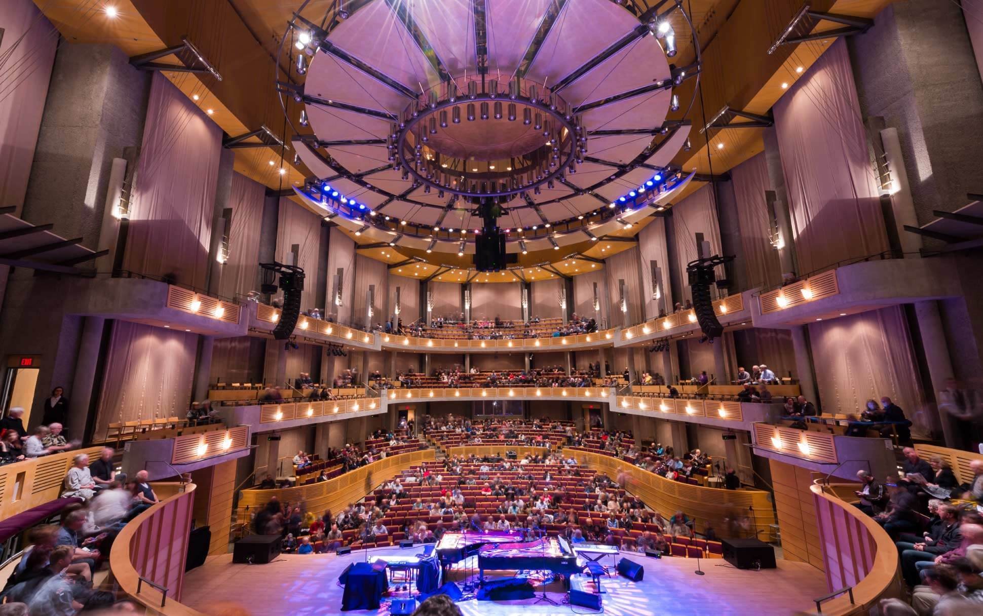 Inside of a large concert hall with a stage and grand piano, surrounded by multiple levels of seating filled with people, and a large circular lighting fixture hanging from the ceiling.