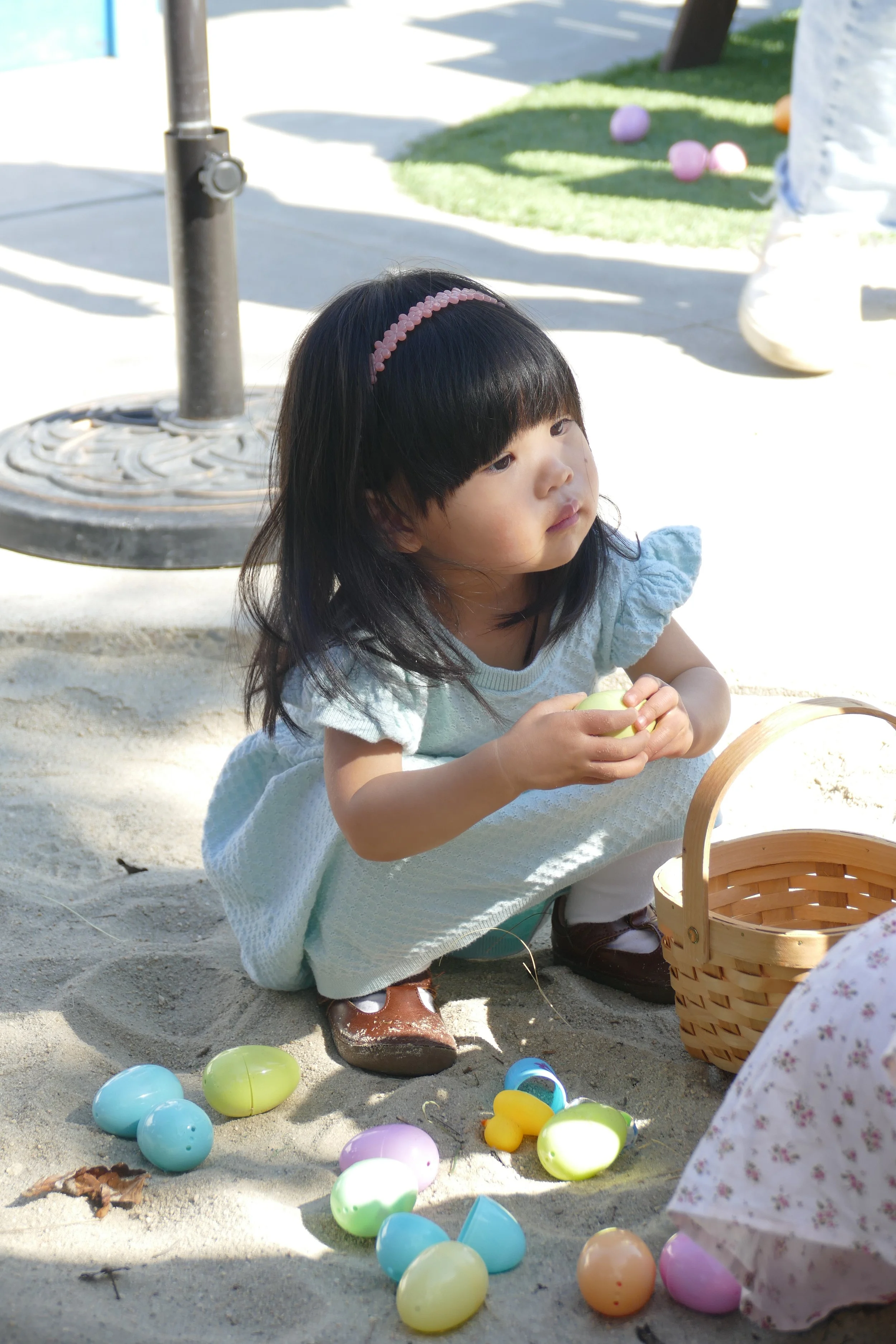 A young girl in a light blue dress with a pink headband is crouching on the ground, holding a yellow plastic egg, surrounded by colorful plastic eggs scattered on sandy ground. There is a wicker basket nearby, and part of another child in a floral dress is visible, with some green grass and colorful objects in the background.