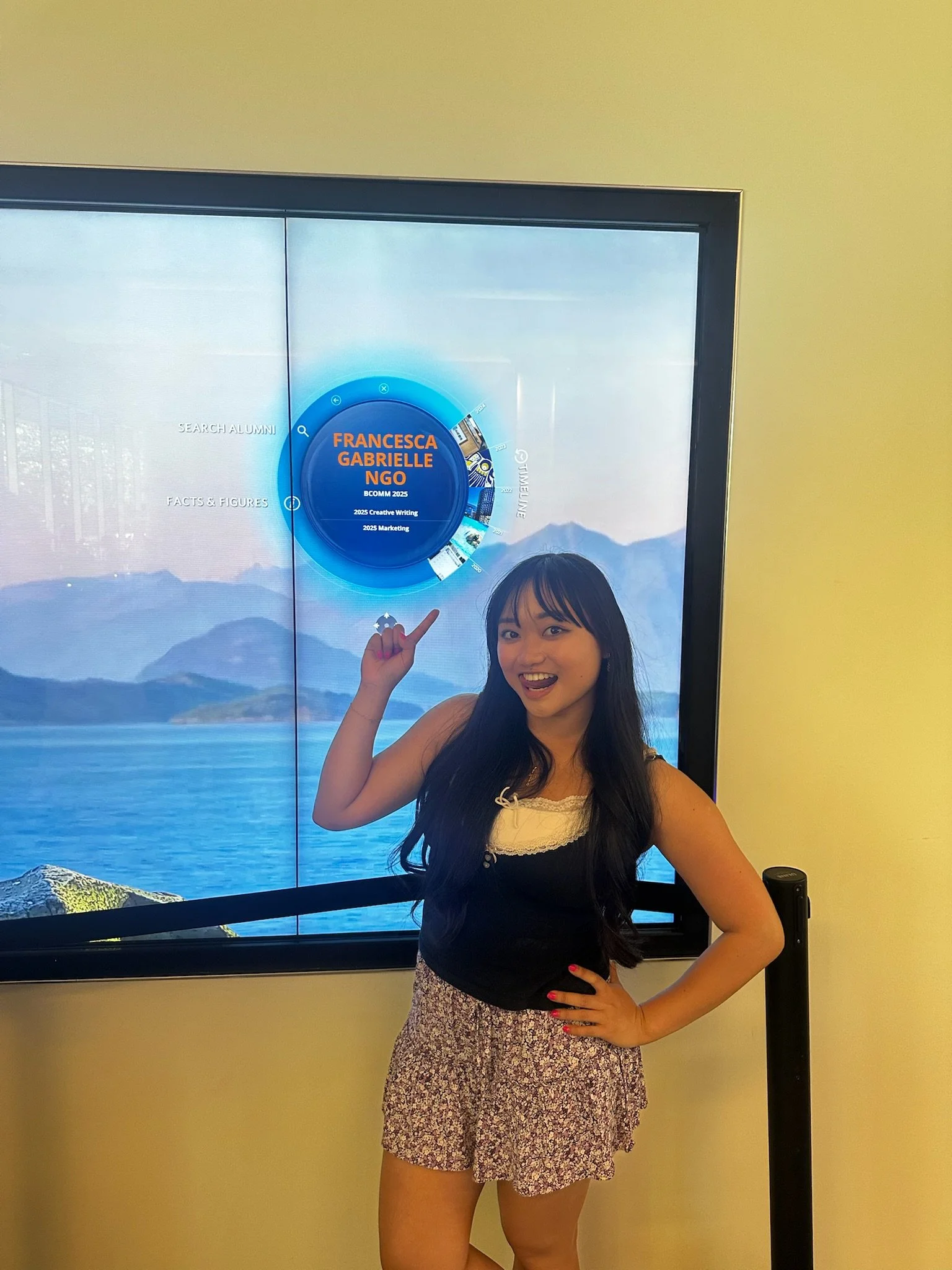 A young woman with long dark hair poses in front of a large digital screen displaying a scenic mountain and lake landscape. She is wearing a black sleeveless top and patterned shorts, smiling and pointing towards the screen with her right hand.