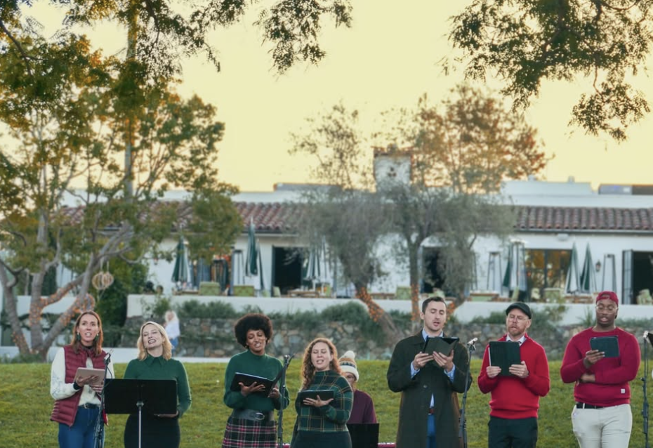 A group of seven diverse people singing outdoors in front of houses and trees during daytime.