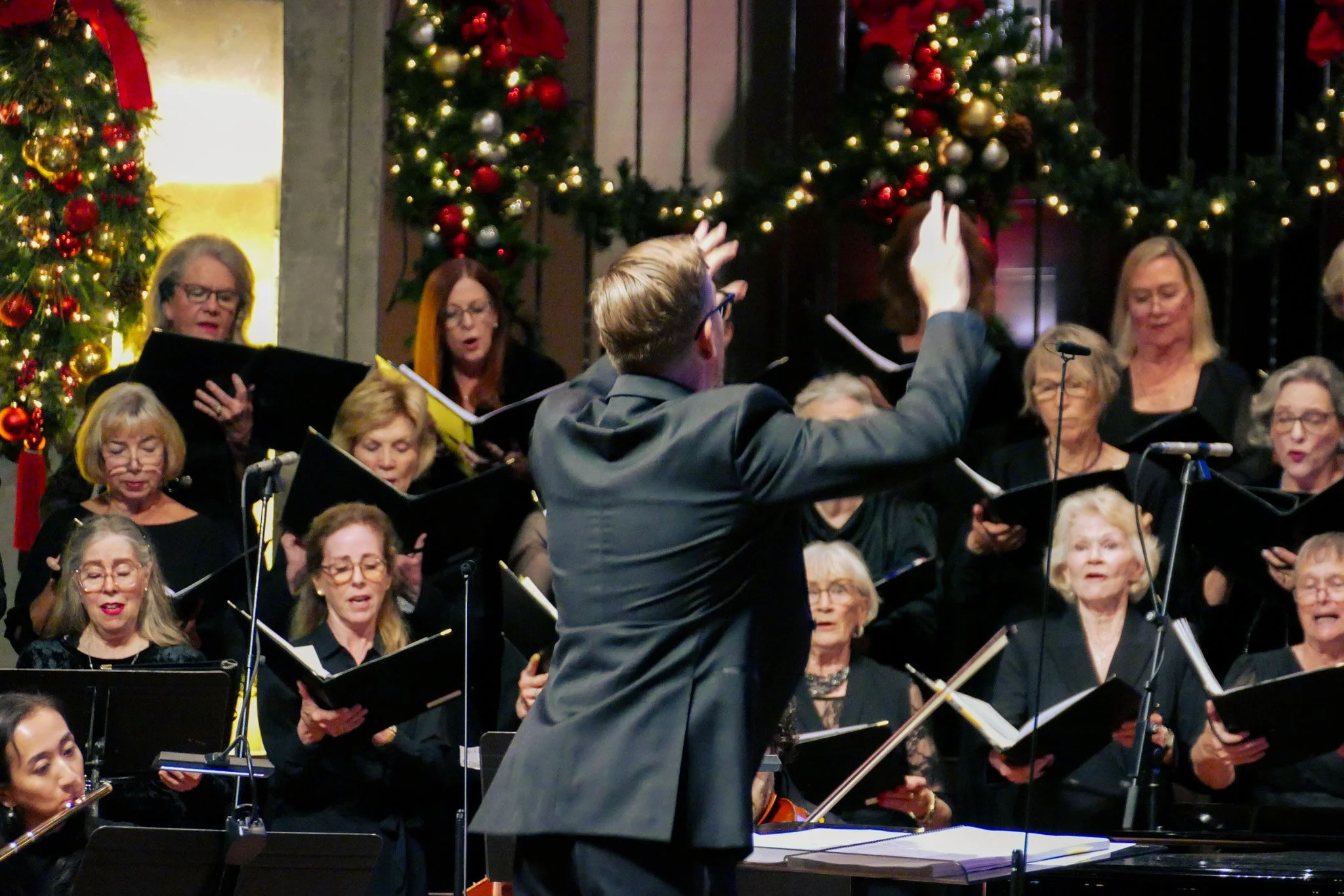 A choir of mostly older women and some men performing in front of Christmas trees decorated with red, gold, and silver ornaments and lights, with a conductor leading them.