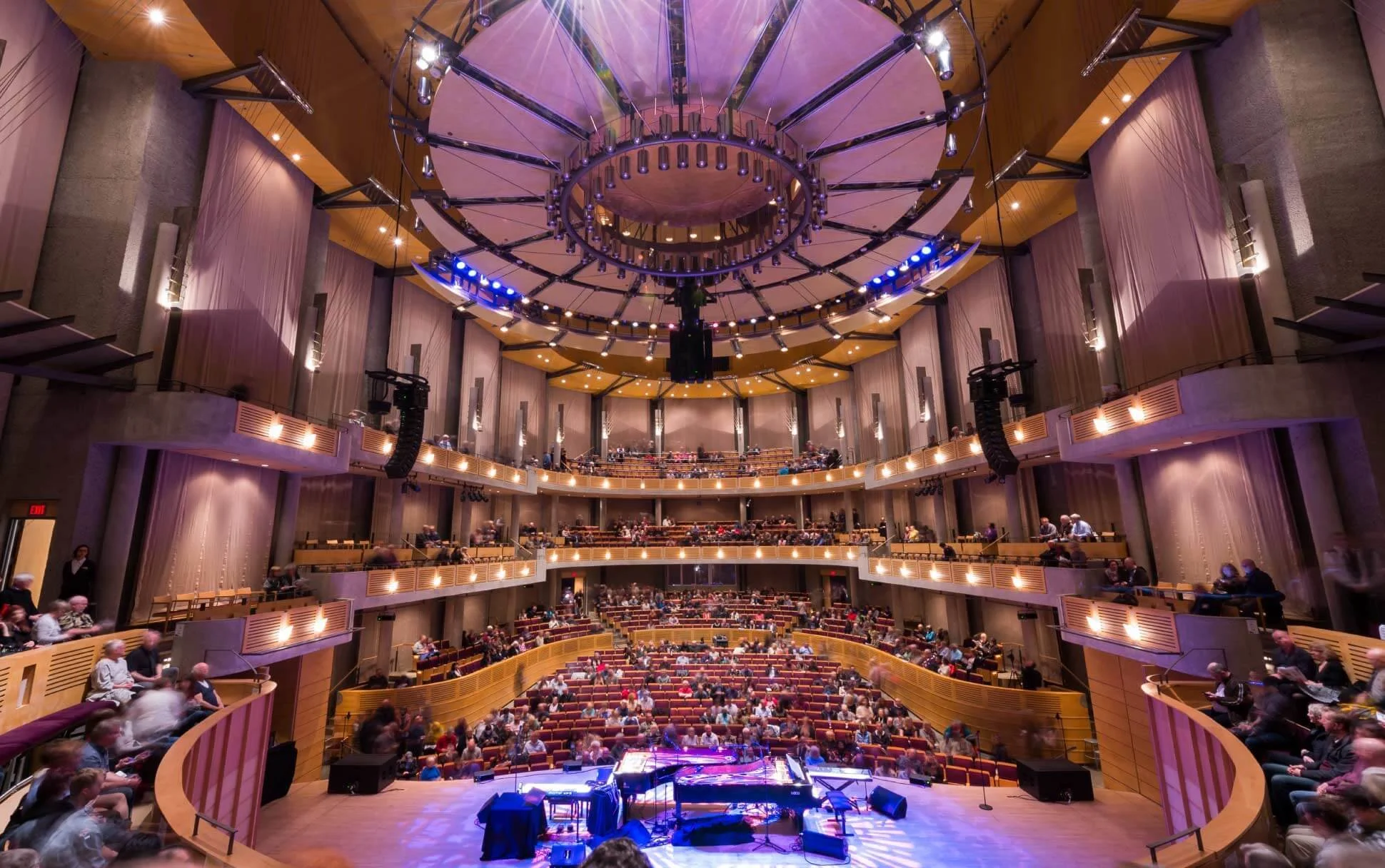 Inside a large concert hall with a grand stage and a grand piano ready for a performance. Audience members are seated in multiple levels of balconies and in the main floor area. The ceiling features an elaborate circular light fixture with blue accent lights.