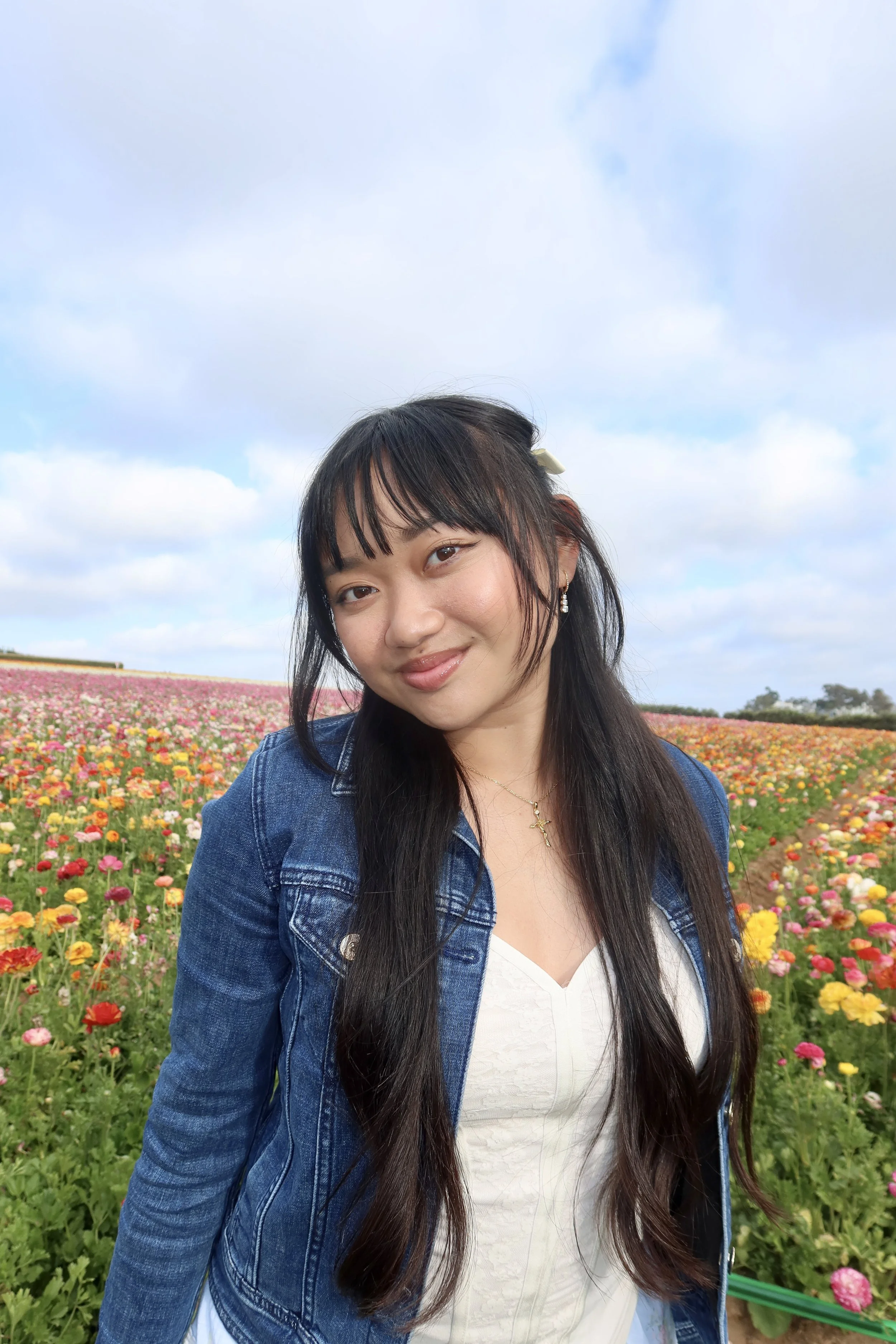 Young woman with long dark hair, wearing a denim jacket, and smiling in a colorful flower field with an overcast sky.