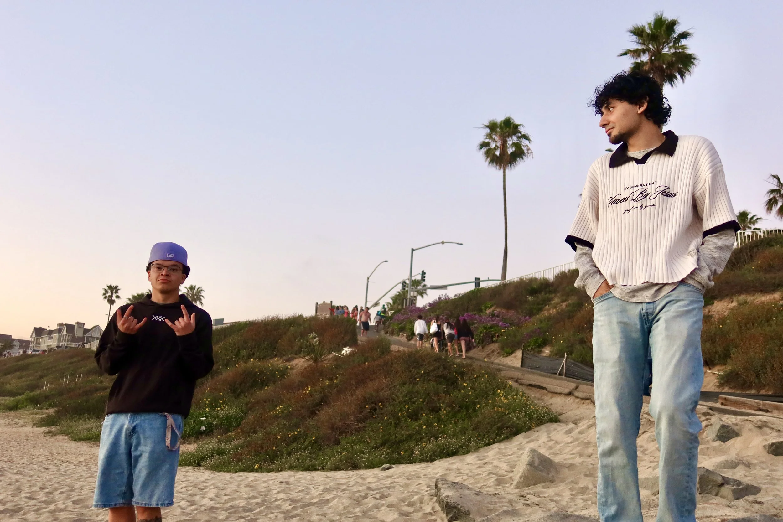 Two young men standing on a sandy beach with palm trees in the background. One man is wearing a gray baseball cap, glasses, and a black hoodie, while the other man has curly black hair and is dressed in a ribbed white and black shirt with jeans. They are outdoors during late afternoon or early evening.