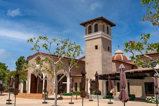 A Spanish-style building with a tower, surrounded by trees and closed umbrellas in an outdoor plaza.