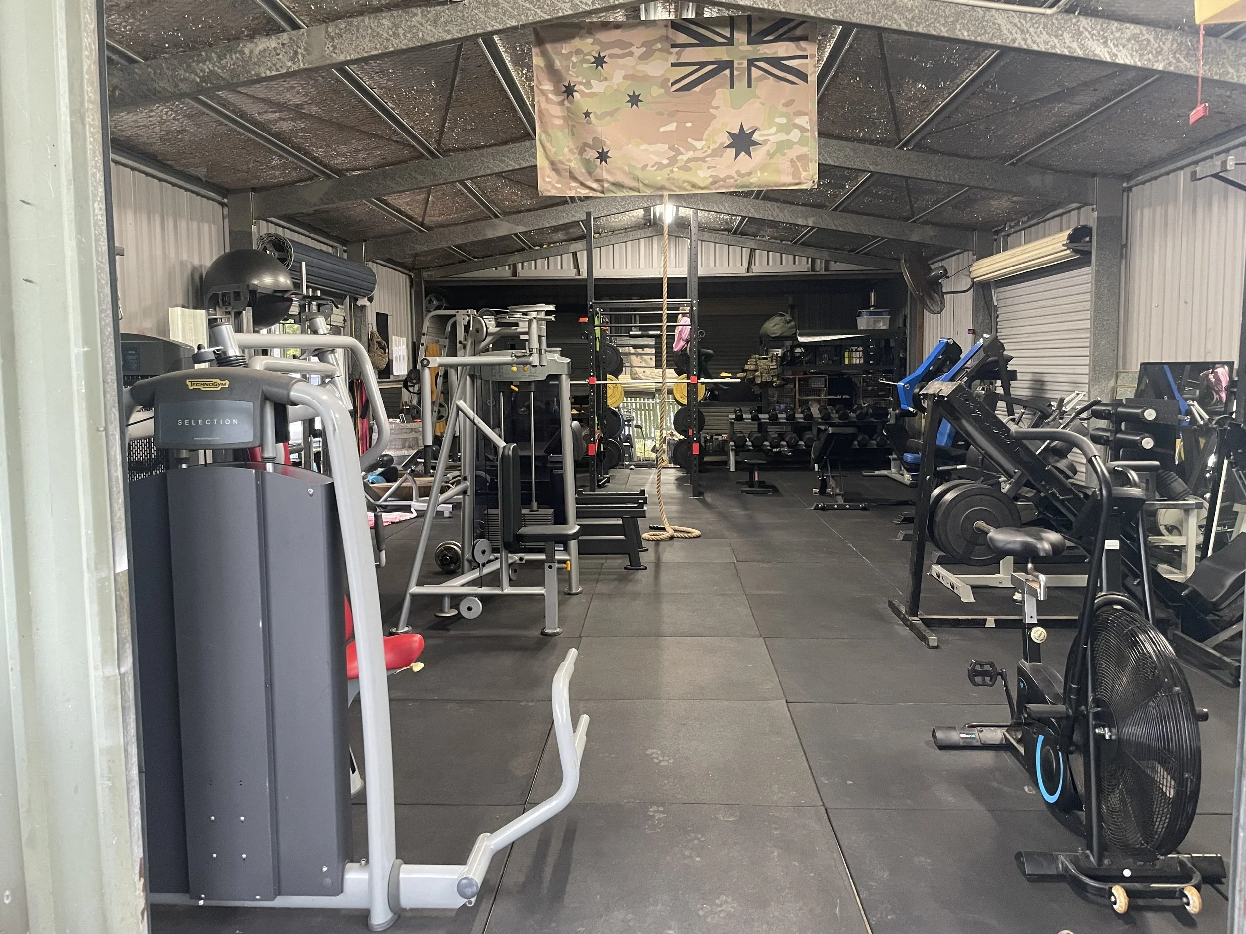 Inside a small garage with various gym equipment including weights, machines, and cardio equipment, with a camouflage flag hanging from the ceiling.