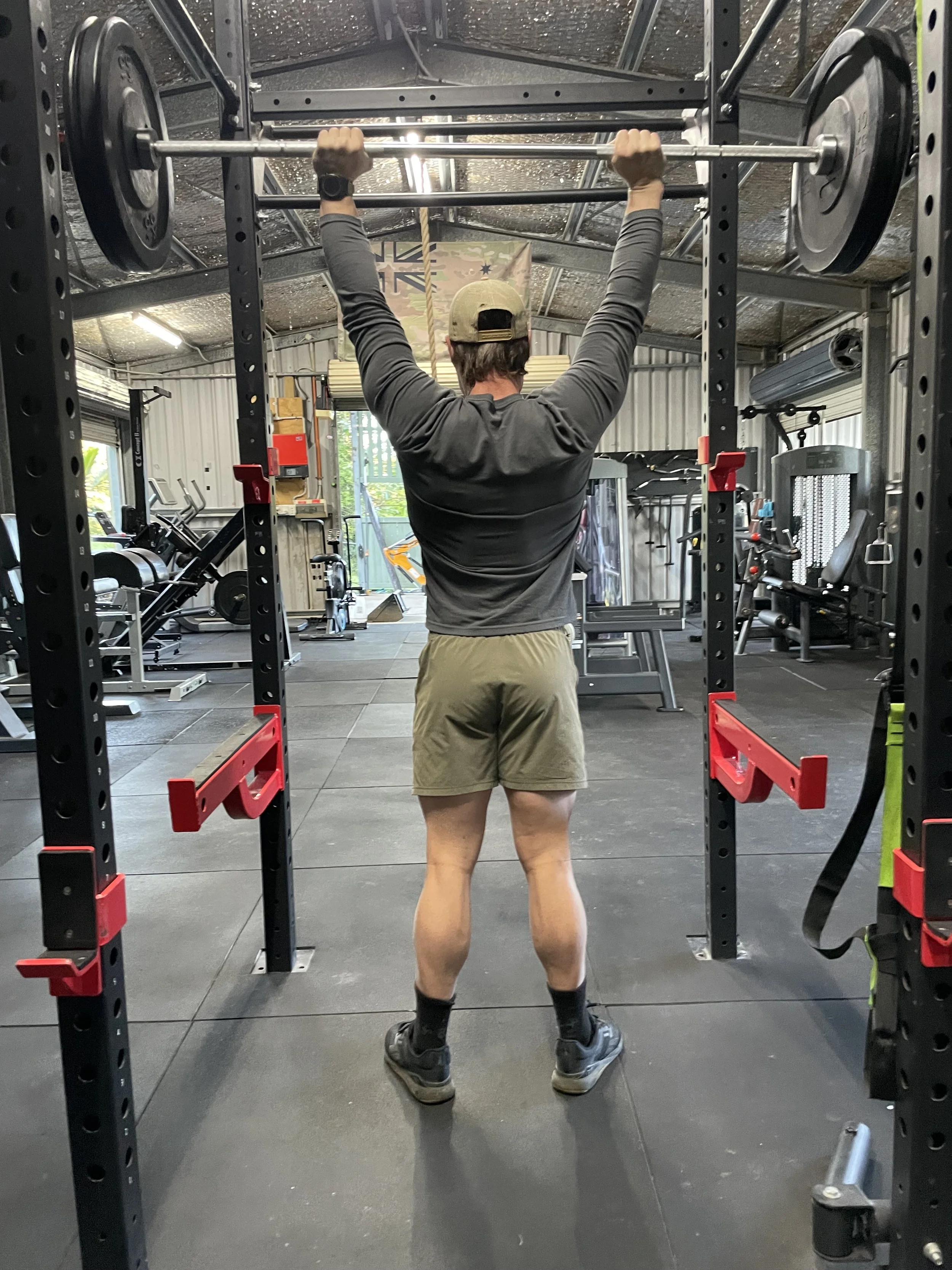 A person wearing a beige cap, gray long-sleeve shirt, tan shorts, black socks, and sneakers is standing inside a gym, lifting a barbell loaded with weight plates above their head. The gym has various workout equipment and is illuminated with overhead lighting.