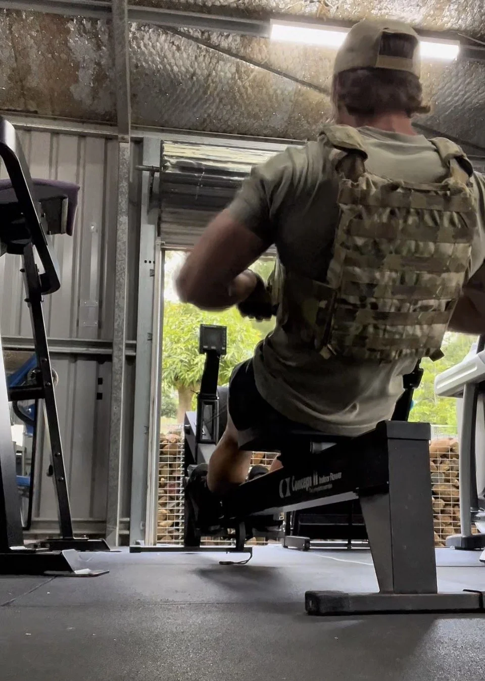 A person wearing a camouflage tactical vest and a baseball cap is running on a treadmill inside a garage with open doors, with greenery visible outside.