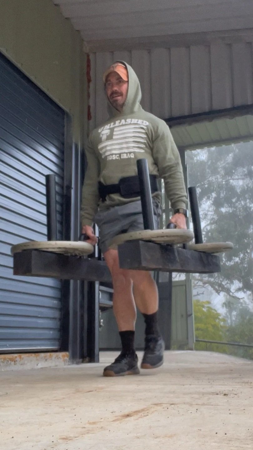 Man carrying two chairs, wearing a hoodie, shorts, and hiking boots, standing on a porch with a closed roller shutter door to his left and trees visible in the background.