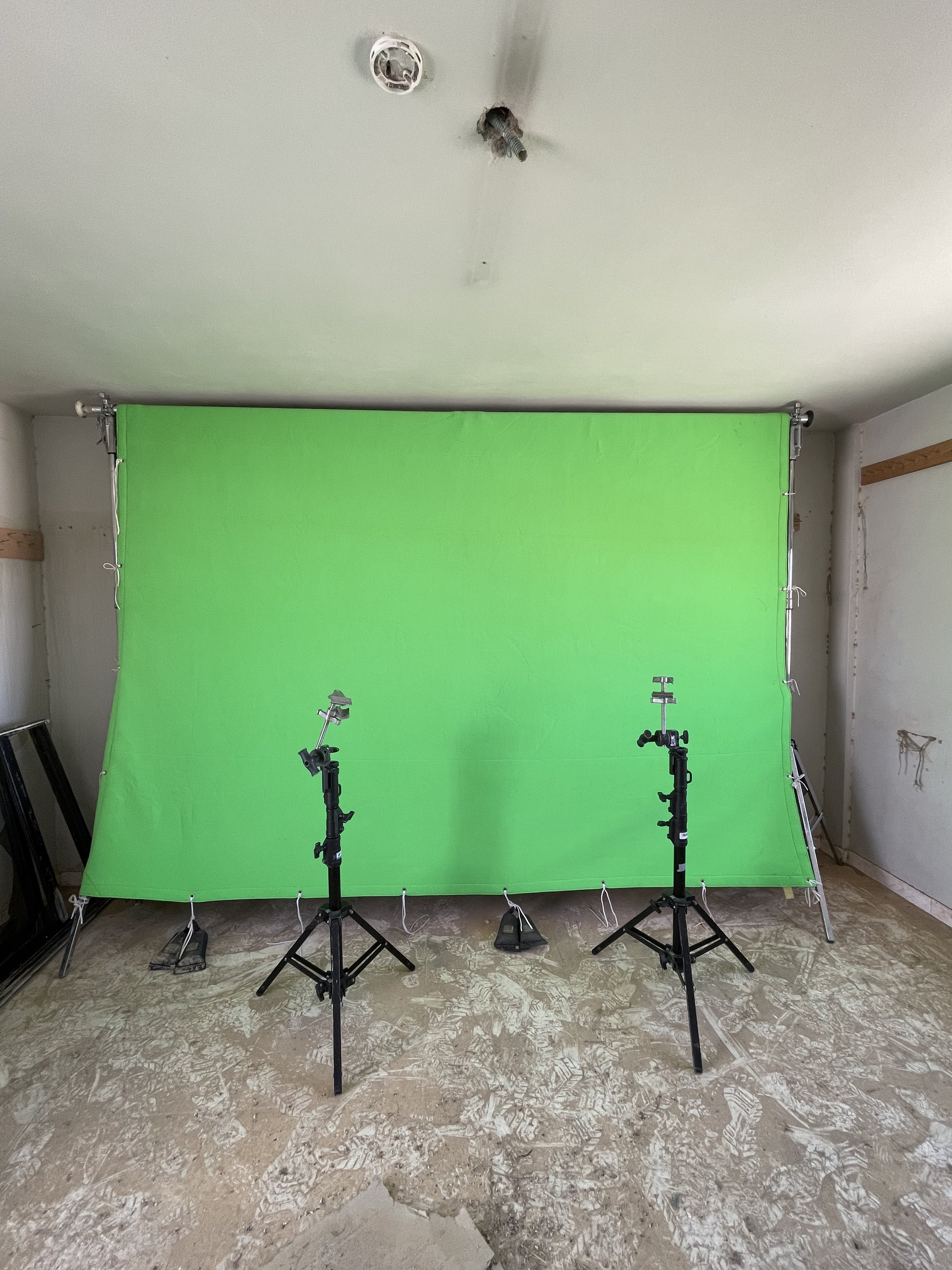 A photography studio setup with a green backdrop, two light stands with lights in front, and a ceiling light fixture. The floor has a pattern with footprints and construction debris.