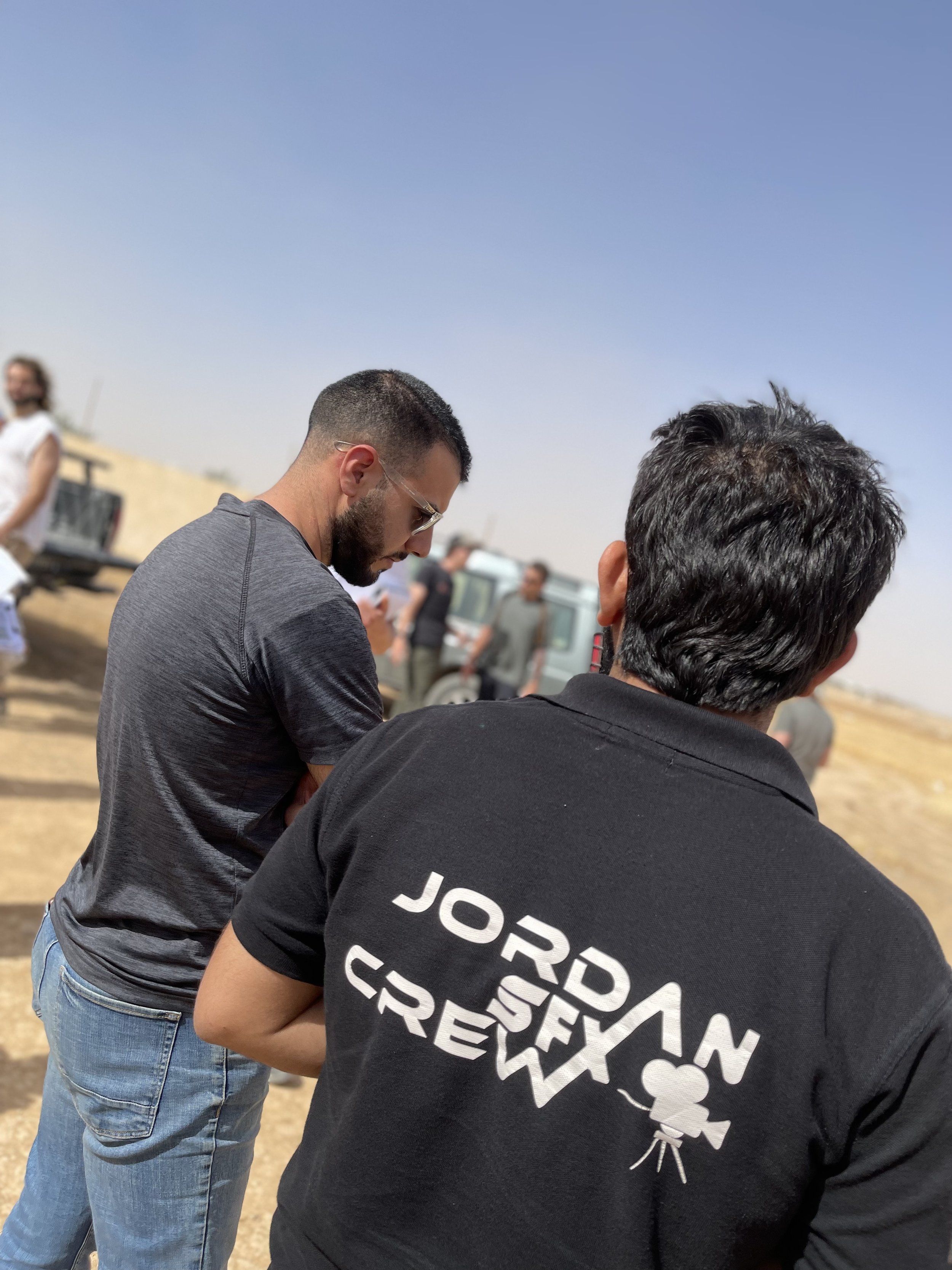 People outdoors, some wearing black and gray casual clothing, with a clear blue sky, a woman in white in the background, and a few other people and vehicles in a dusty environment.