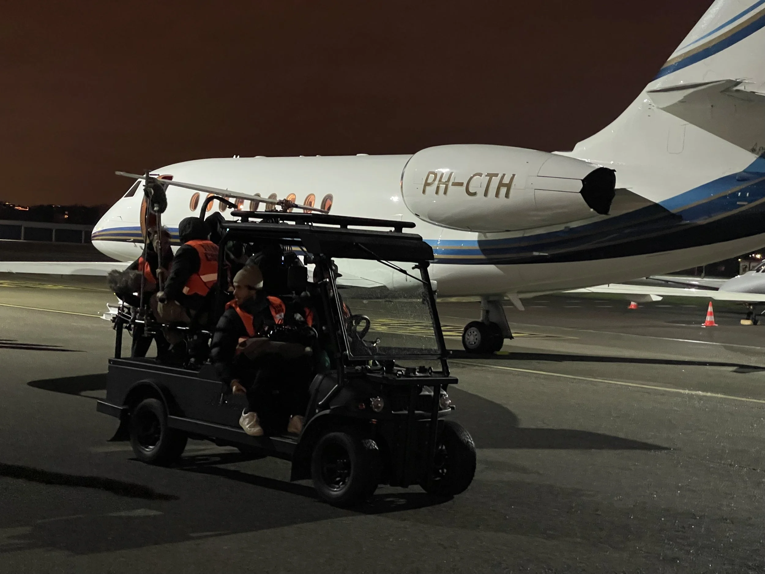 Nighttime scene on an airport tarmac showing a small jet airplane with the registration PH-CTH and a golf cart with two people in orange vests near the jet's nose.