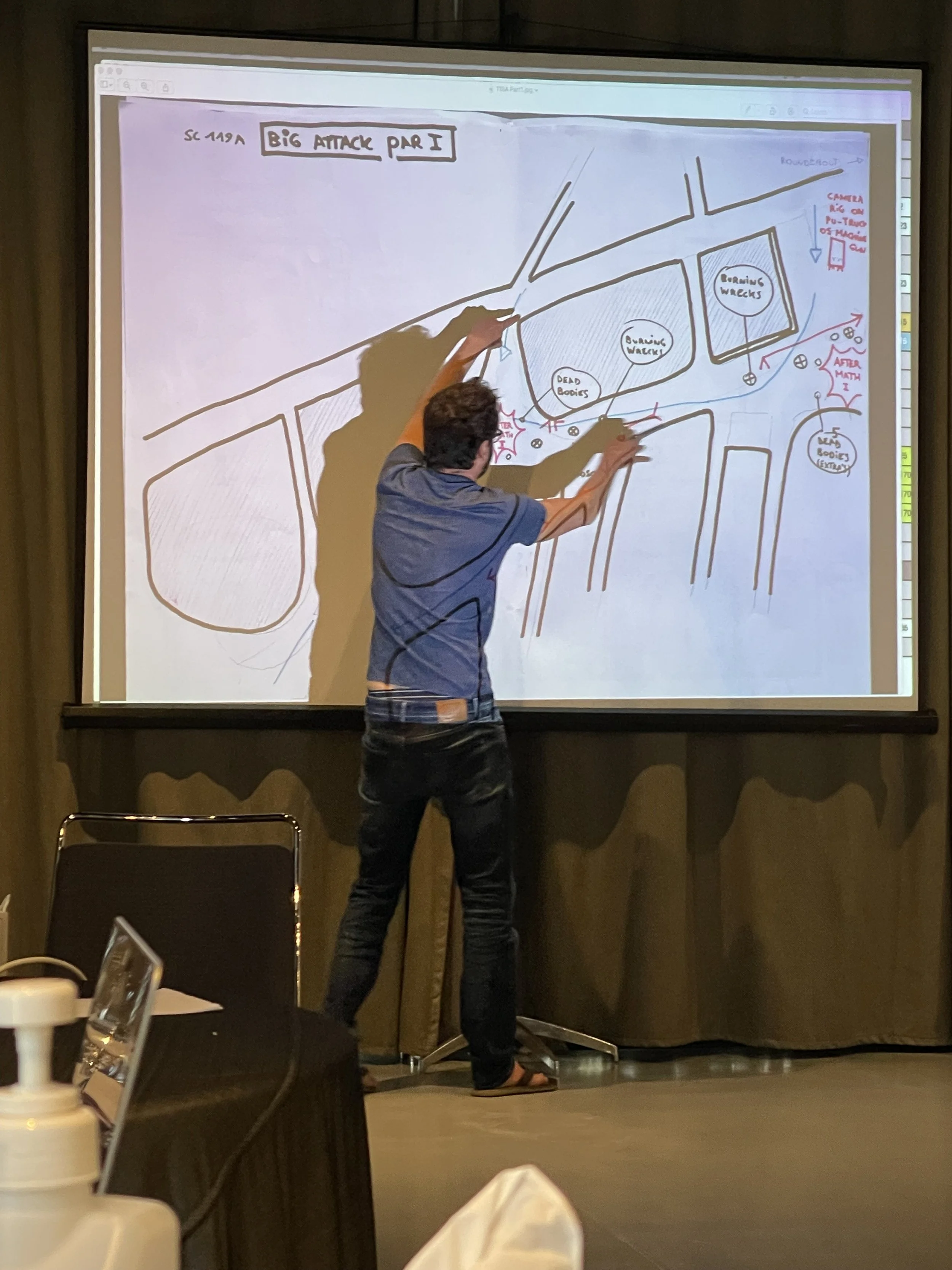 A man standing in front of a large projection screen, pointing at a hand-drawn map or diagram. The map includes handwritten notes and labels related to a large attack scenario, with sections labeled 'Dead Bodies' and 'Bombing Wrecks'. He appears to b