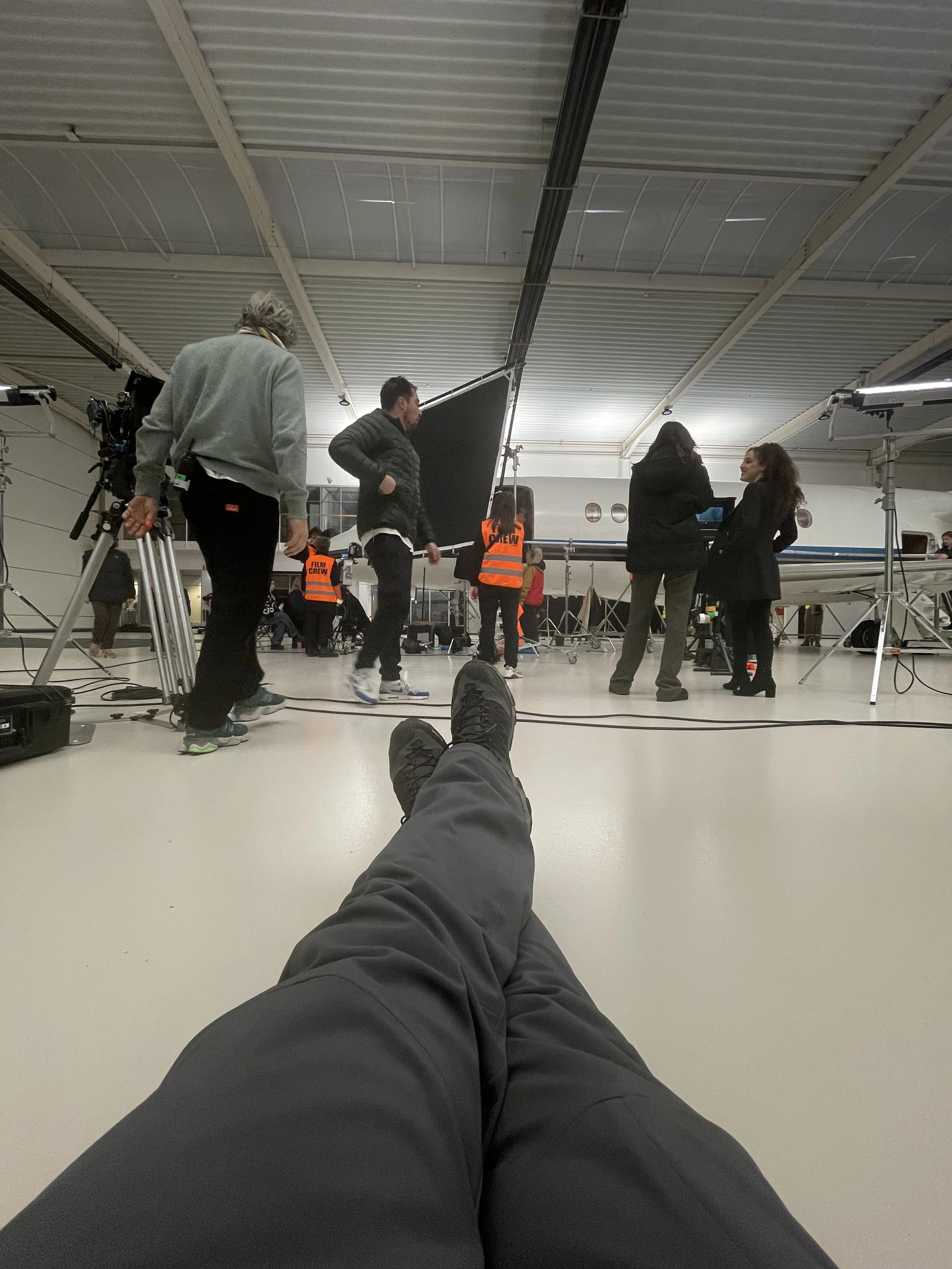 View from a person's perspective, sitting on the floor with legs stretched out, at an airport or similar facility, surrounded by crew members and production equipment, with a large aircraft in the background.